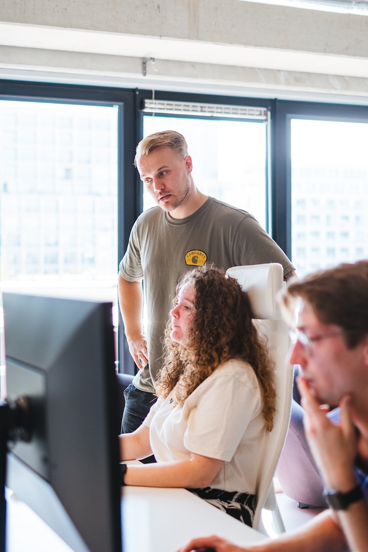 A man stands behind a woman seated at a desk, both focused on a computer screen. Another person sits nearby, slightly out of focus, all appearing to work together in a modern office with large windows.