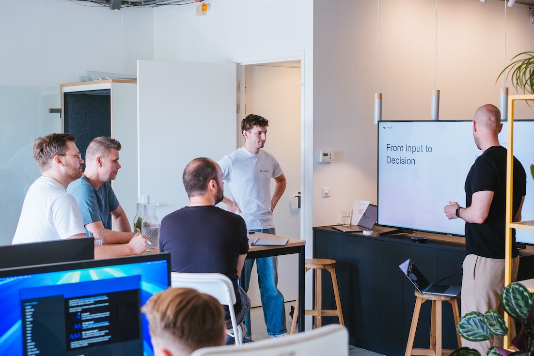 A group of five people in a modern office listen to a presentation. One person stands by a large screen displaying the text "From Input to Decision" while the others are seated, watching attentively.