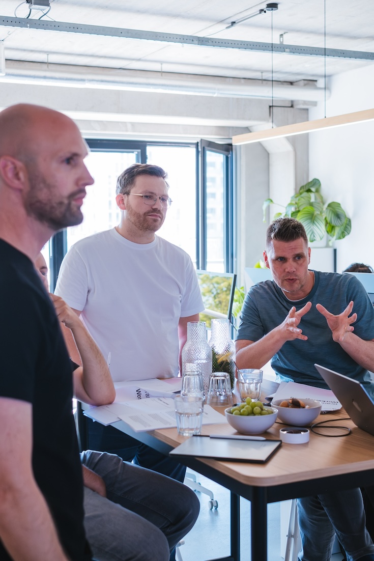 Four men are gathered around a table in a modern office, engaged in discussion. Papers, snacks, drinks, and a laptop are on the table. Large windows and a plant are in the background, letting in natural light.