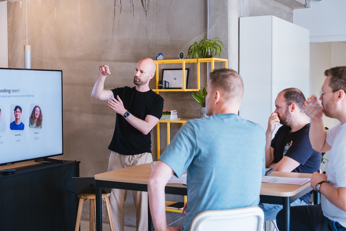 A man stands and gestures while giving a presentation to three seated people in a modern office. A screen displays a “Founding team” slide with three profile photos and names. The audience listens attentively.