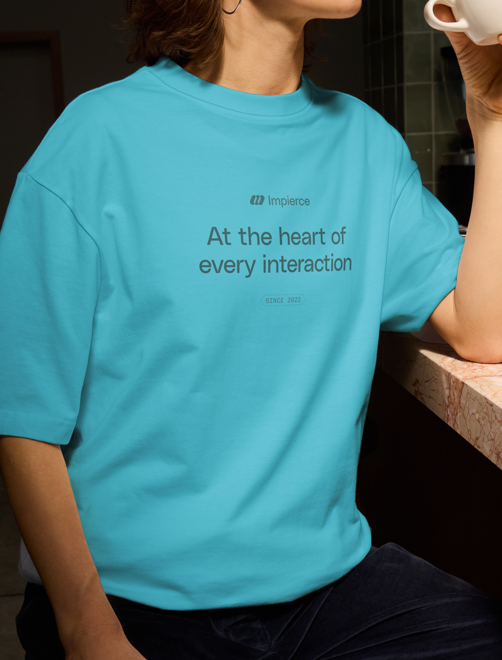 A person in a bright blue t-shirt sits at a counter, holding a white cup. The t-shirt reads, "Impierce At the heart of every interaction SINCE 2022.