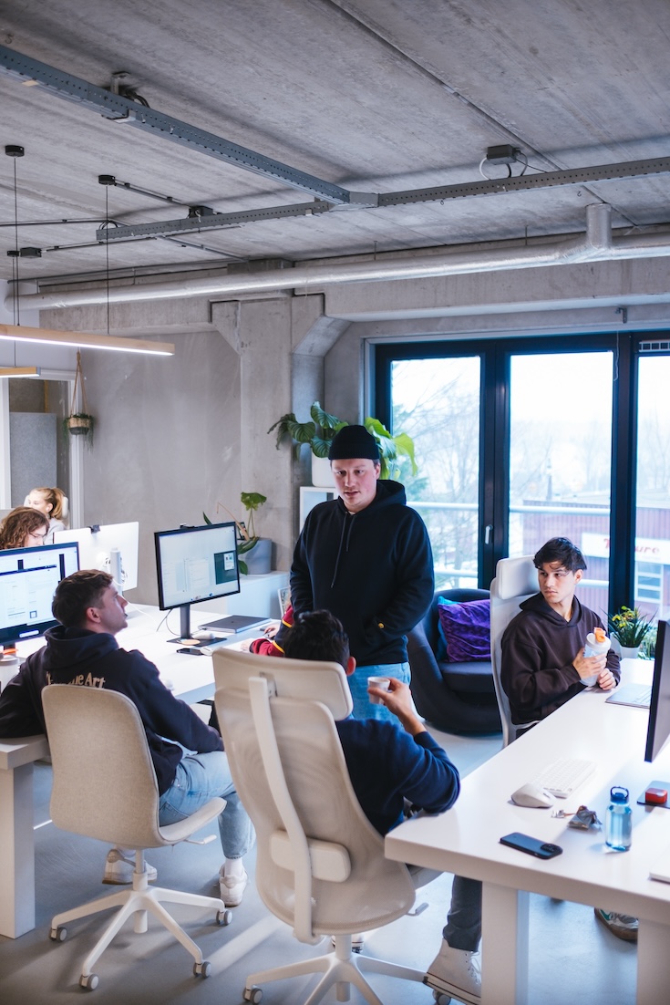 Four young adults are gathered around white desks with computers in a modern office. One person stands while the others sit, engaged in conversation. Large windows and plants add natural light and greenery to the workspace.