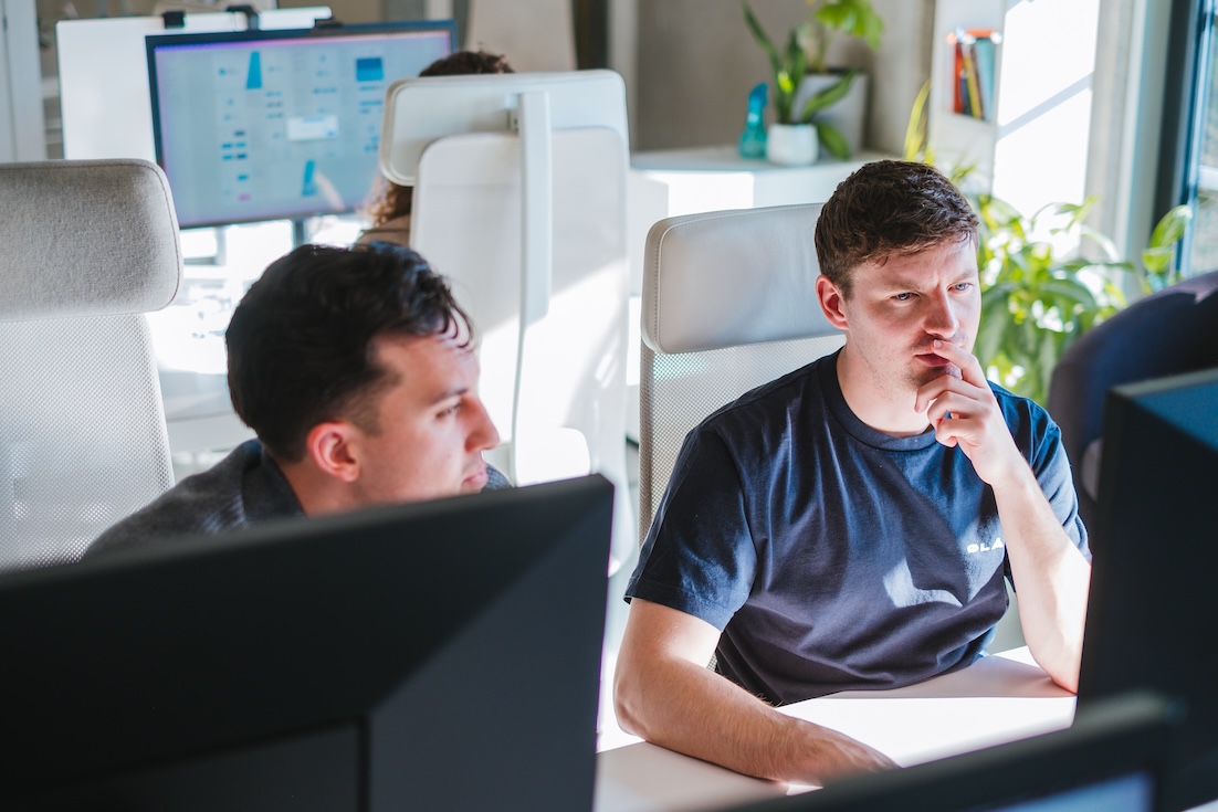 Two men sit at desks in a bright office, focused on computer screens. One man rests his hand on his chin, appearing thoughtful. Office plants and monitors are visible in the background.