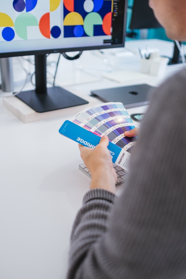 A person holding a Pantone color swatch book at a desk, reviewing color options. A computer monitor displays colorful geometric shapes in the background.