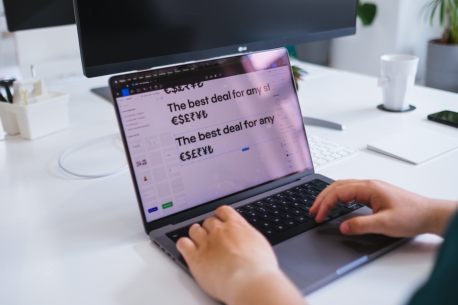 A person types on a laptop at a white desk displaying text with different currency symbols on the screen. Office supplies, a monitor, and a cup are visible in the background.