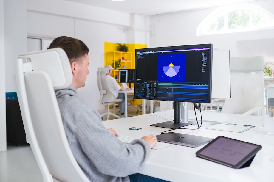 A person sitting at a white desk in a modern office, working on graphic design or animation software displayed on a large monitor. A tablet and keyboard are on the desk, with bright shelves in the background.