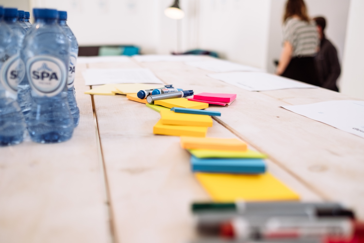 A close-up of a wooden table with colorful sticky notes, markers, papers, and bottles of water. In the background, two people are standing and talking in a bright, modern room.