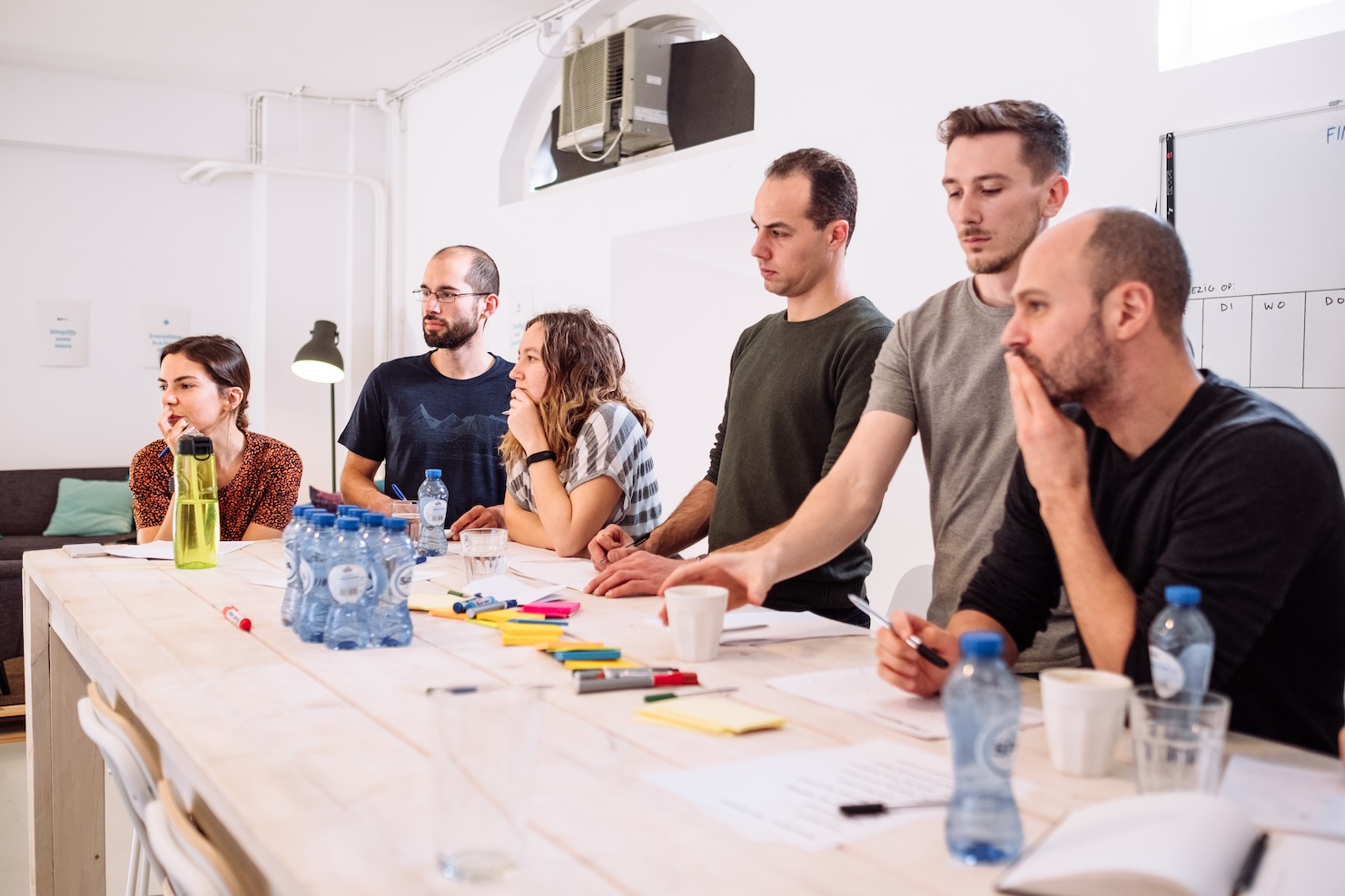 Six people stand around a table covered with water bottles, sticky notes, markers, and papers, appearing focused and engaged in discussion or a meeting in a bright, modern office setting.