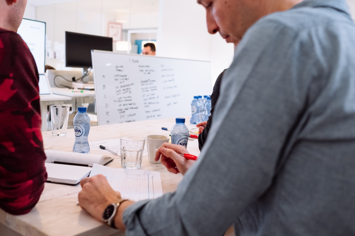 Two people sit at a table with papers and water bottles, one writing with a red pen. A whiteboard with notes and a computer monitor are visible in the background of a bright office setting.
