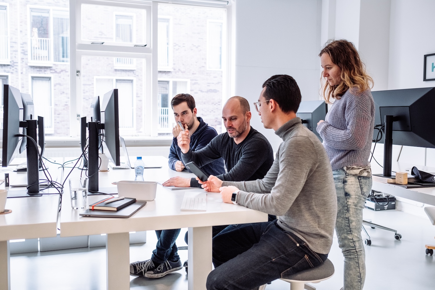 Four people in casual clothing meet around a table with computers in a bright office. Two are seated, discussing papers and a tablet, while two others stand nearby listening. Large windows and monitors are in the background.