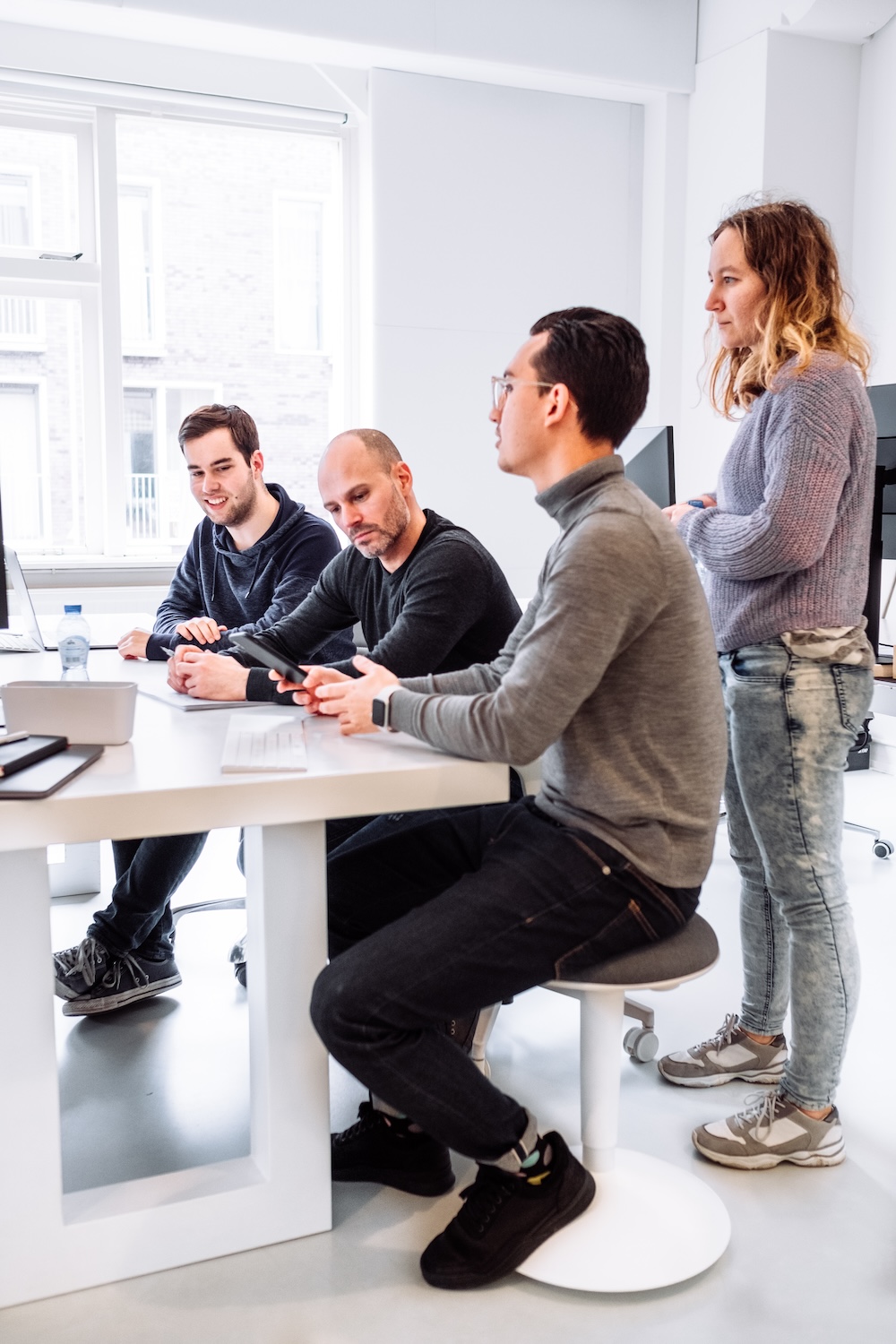 Four people are working together in a bright office. Three are seated at a table with laptops, while one person stands nearby. They appear engaged and focused in a collaborative discussion.