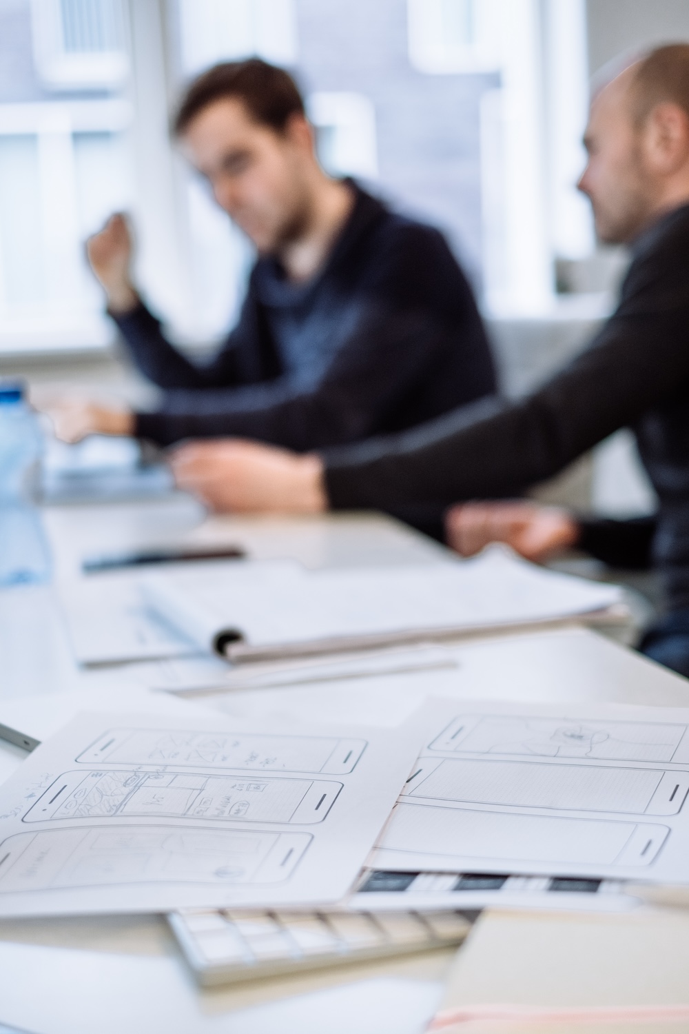 Two people sit at a table with laptops and notebooks, discussing work. In the foreground, there are printed comic or storyboard sketches in focus, while the people are blurred in the background.