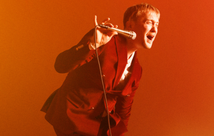 Alexander Lövmark Trio sjunger iklädd röd kostym med orange/röd bakgrund