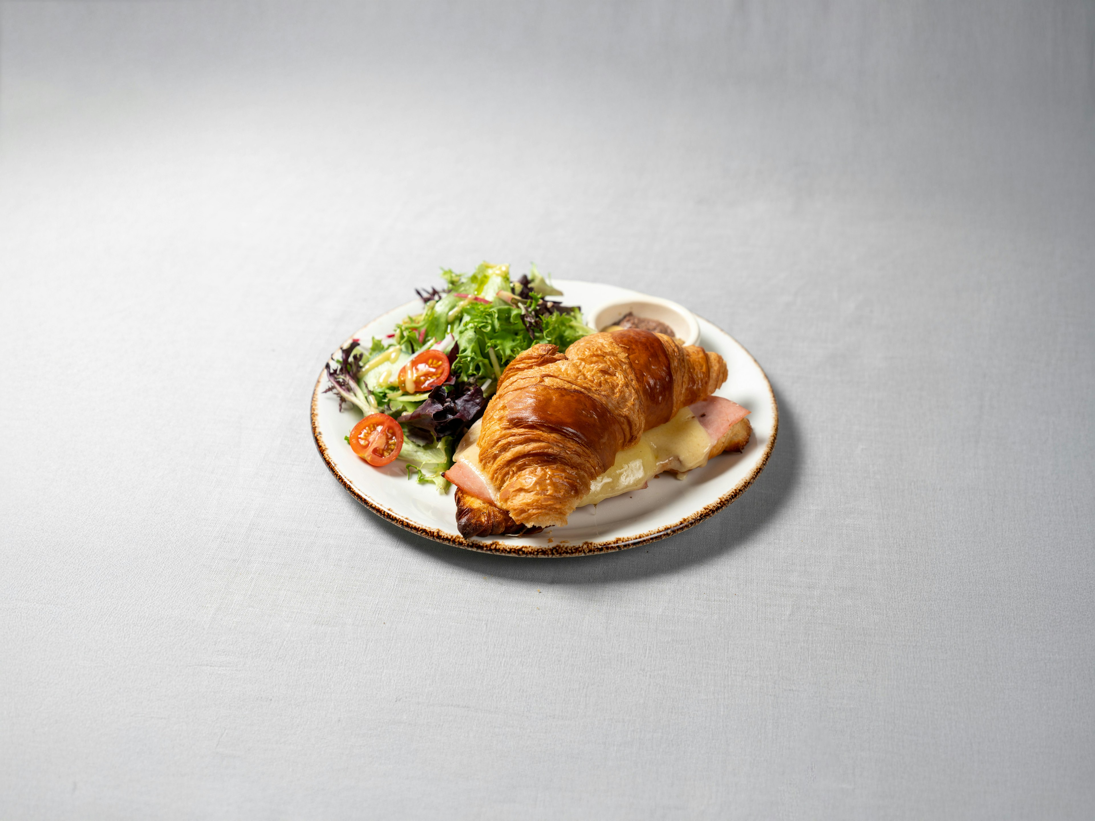 A plate with a ham and cheese croissant, a small green salad with cherry tomatoes, and a small cup of dressing, set on a light gray surface.