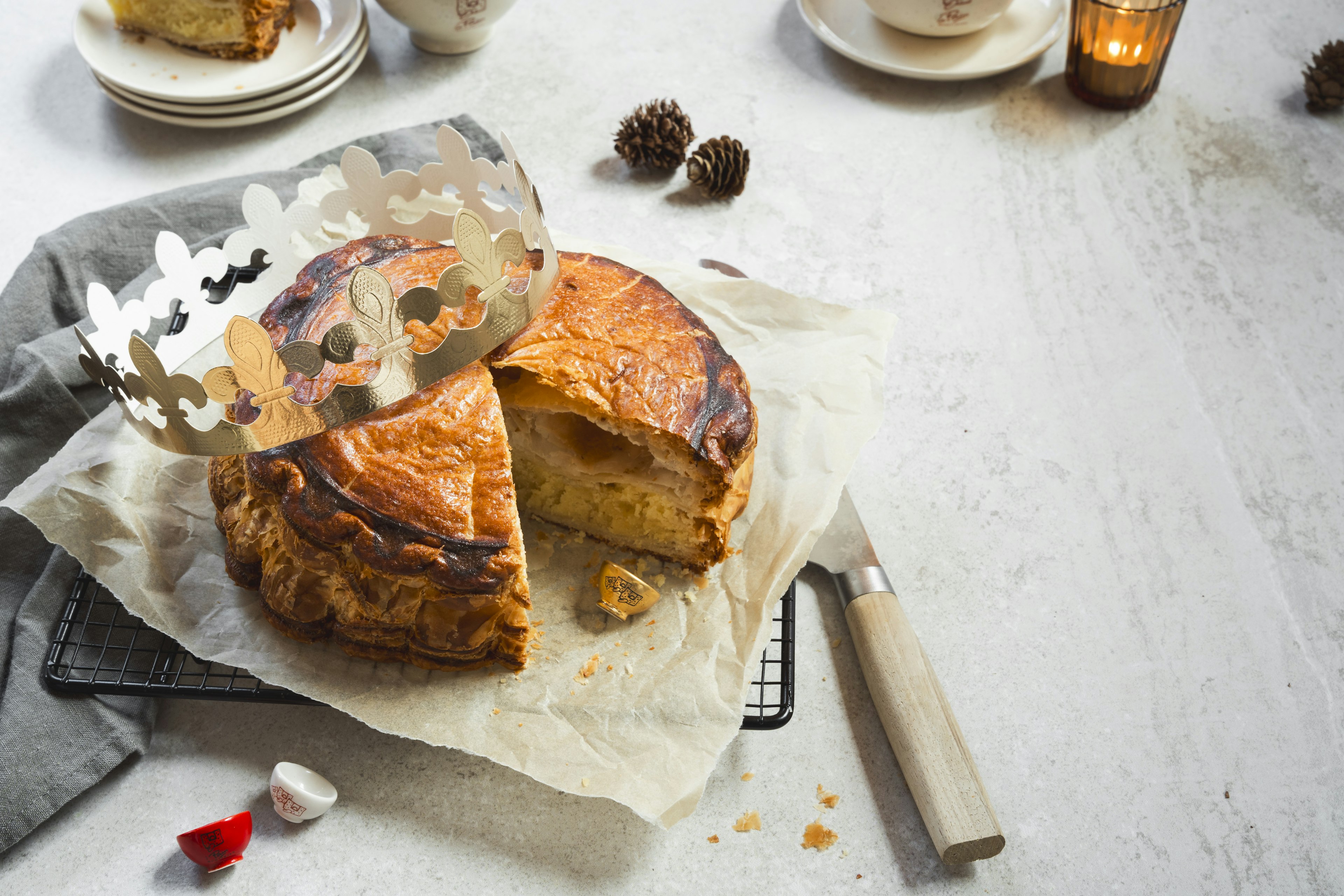 Een goudbruine galette des rois ligt op perkamentpapier, deels gesneden, met een decoratieve papieren kroon erop. Een mes ligt vlakbij en kleine beeldjes en dennenappels liggen verspreid over het lichte oppervlak.