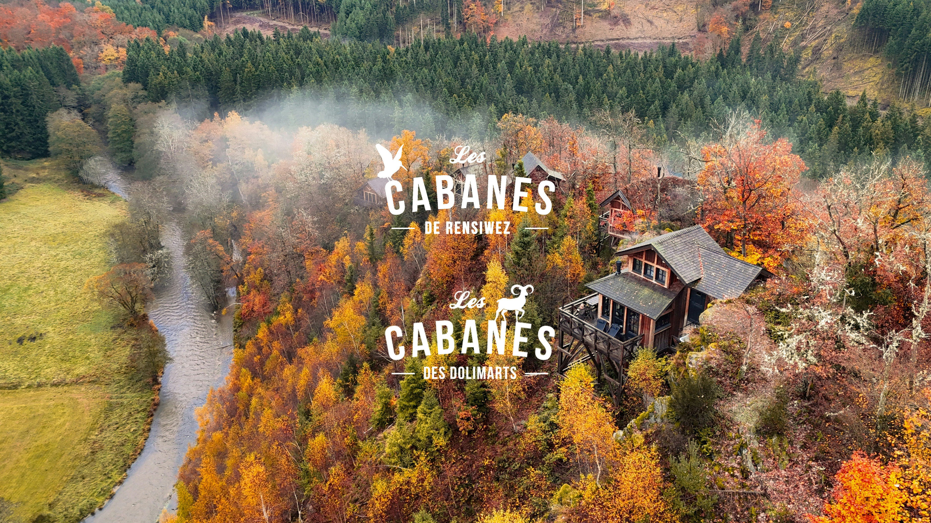 Luchtfoto van hutten genesteld tussen levendige herfstbomen op een heuvel bij een kronkelende rivier, met bos op de achtergrond. De tekst Les Cabanes de Rensiwez en Les Cabanes des Dolimarts is overlaid.