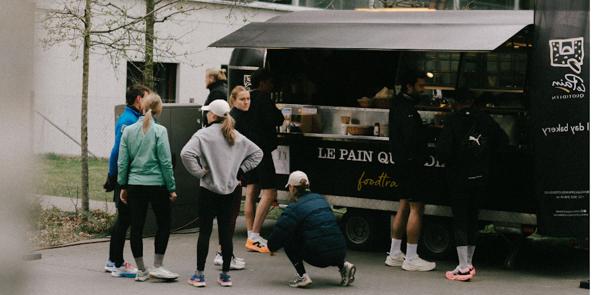 A group of people in athletic clothing stand and crouch near a black food truck labeled Le Pain Quotidien on a paved walkway, with trees and a building visible in the background.