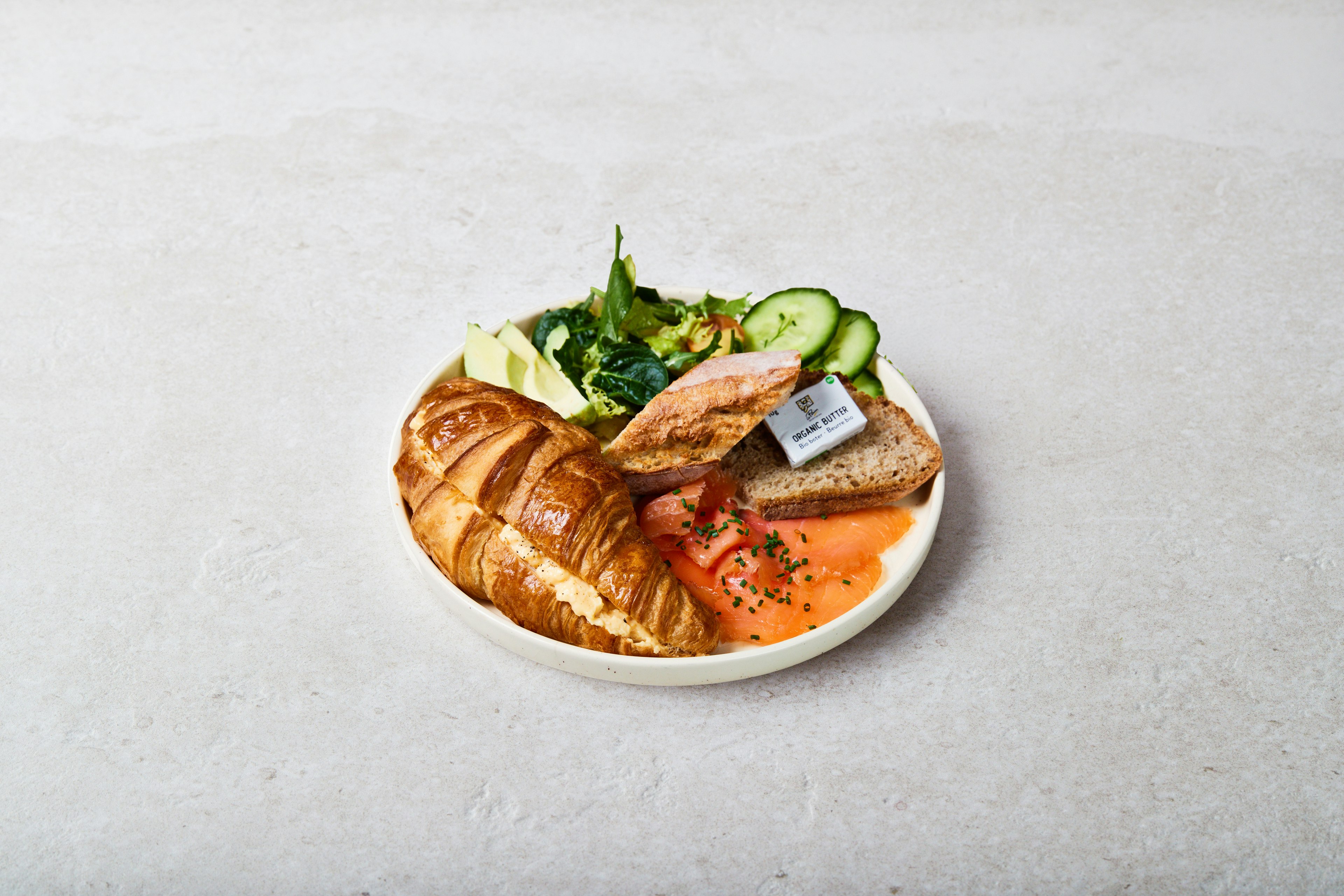 A plate with a croissant, smoked salmon topped with herbs, sliced avocado, cucumber, salad greens, grilled bread, and a small container of butter on a light background.