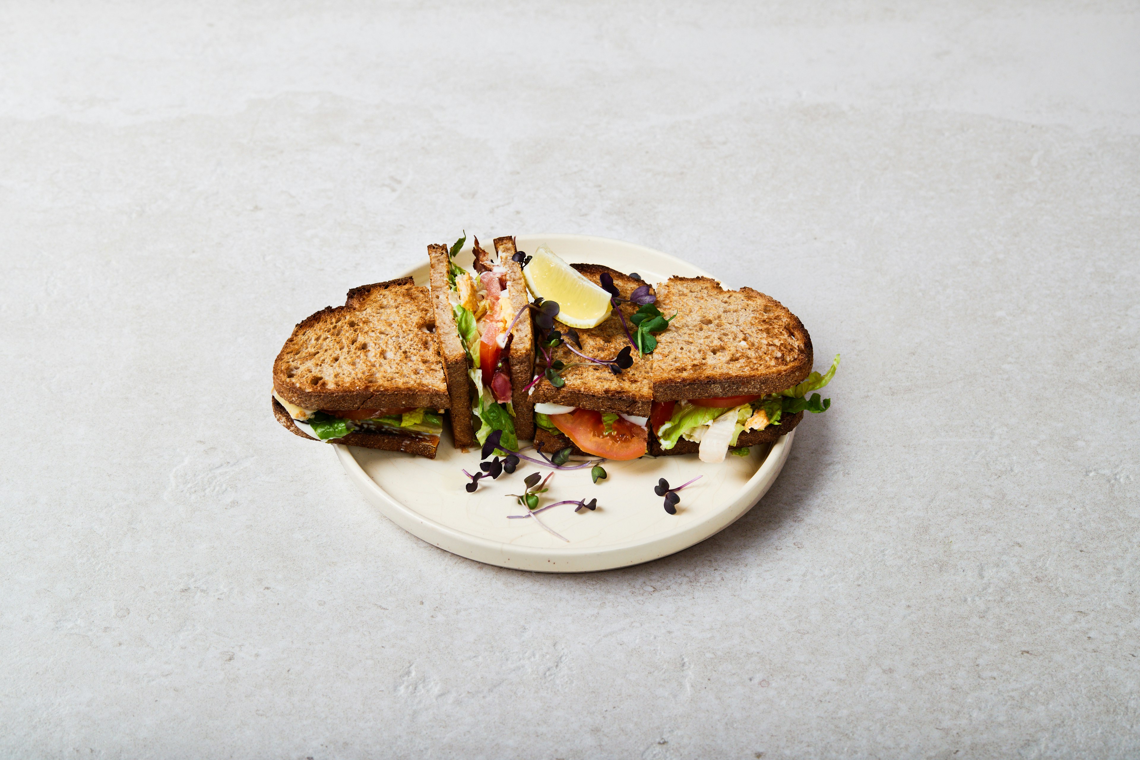 A sandwich with whole grain bread, lettuce, tomato, and other fresh ingredients is cut in half and served on a white plate, garnished with microgreens and a lemon wedge.