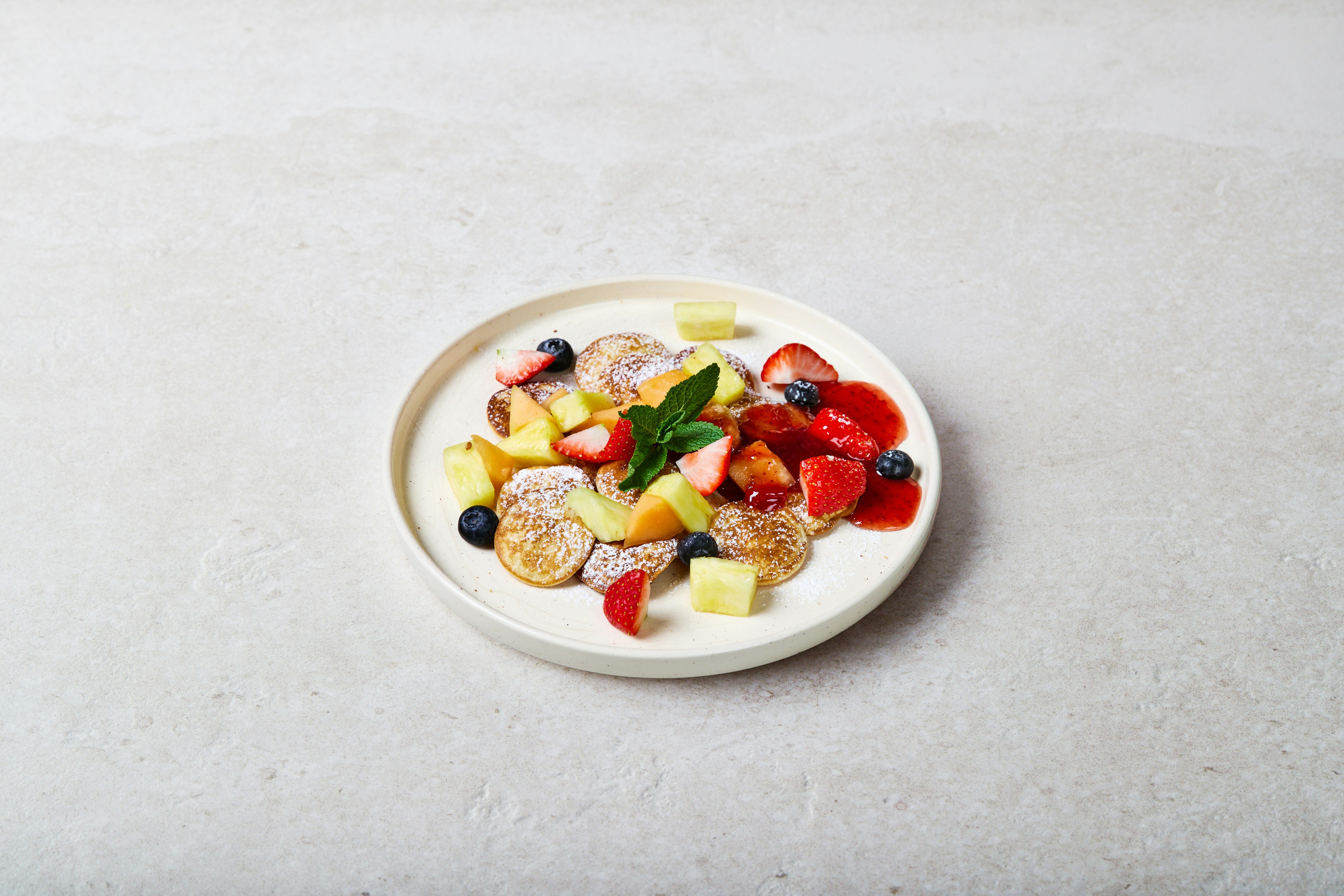 A plate of mini pancakes topped with powdered sugar, fresh strawberries, blueberries, pineapple chunks, and a sprig of mint, on a light-colored background.