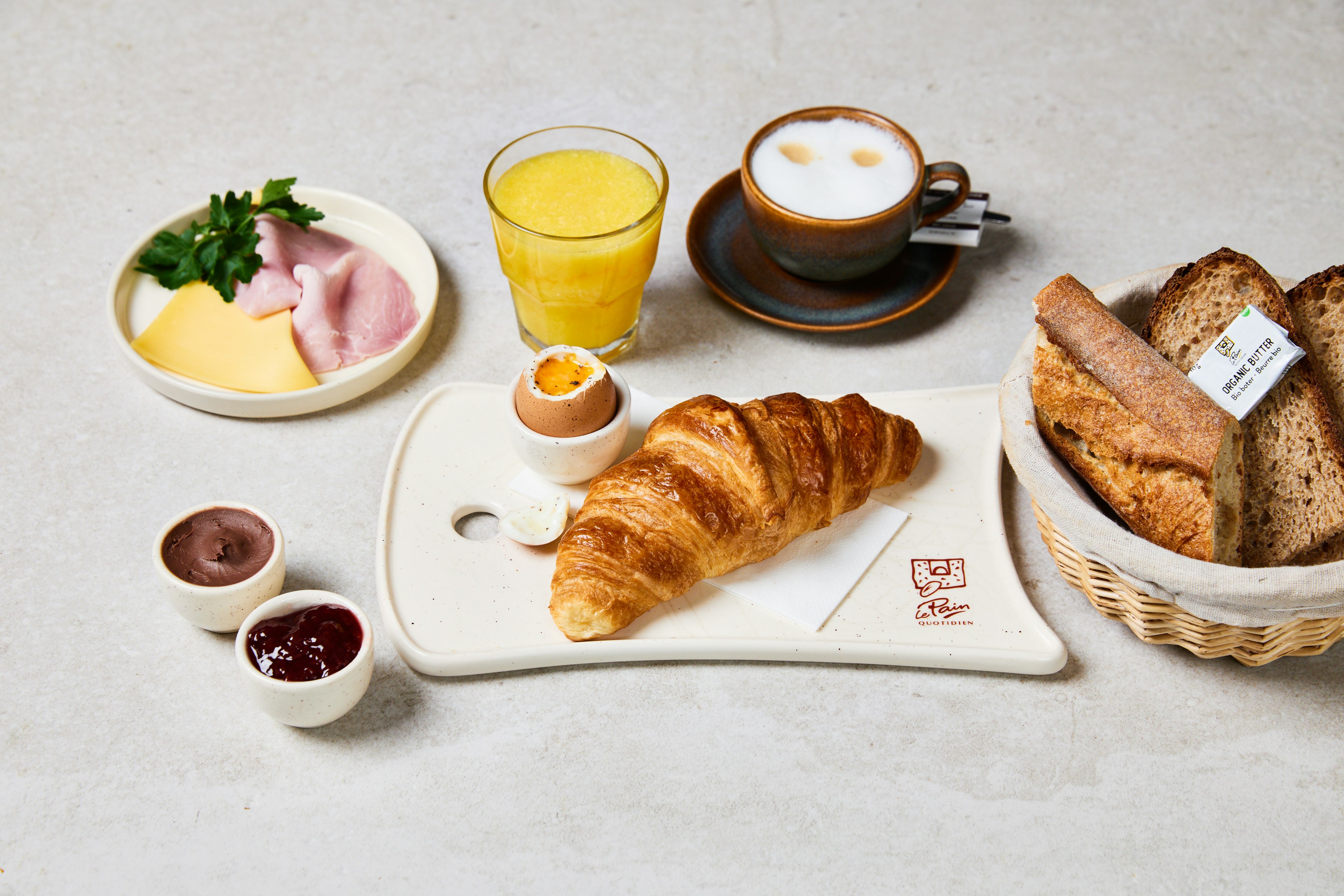 A breakfast spread with a croissant, soft-boiled egg, slices of ham and cheese, orange juice, cappuccino, bread basket, and small bowls of chocolate spread and jam on a light surface.