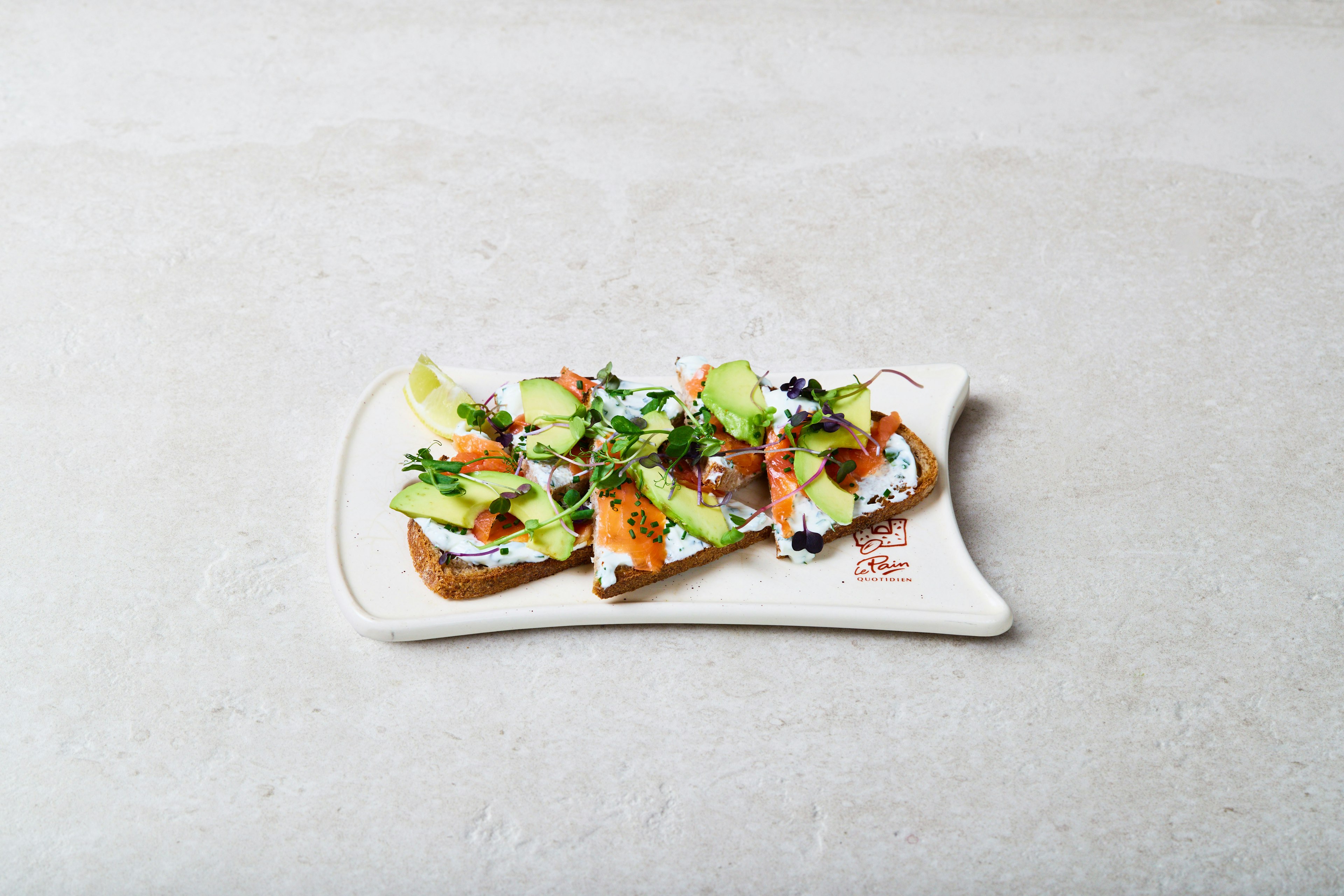 Open-faced sandwich topped with smoked salmon, avocado slices, microgreens, herbs, and lemon wedges on a rectangular white plate, set on a light-colored textured surface.