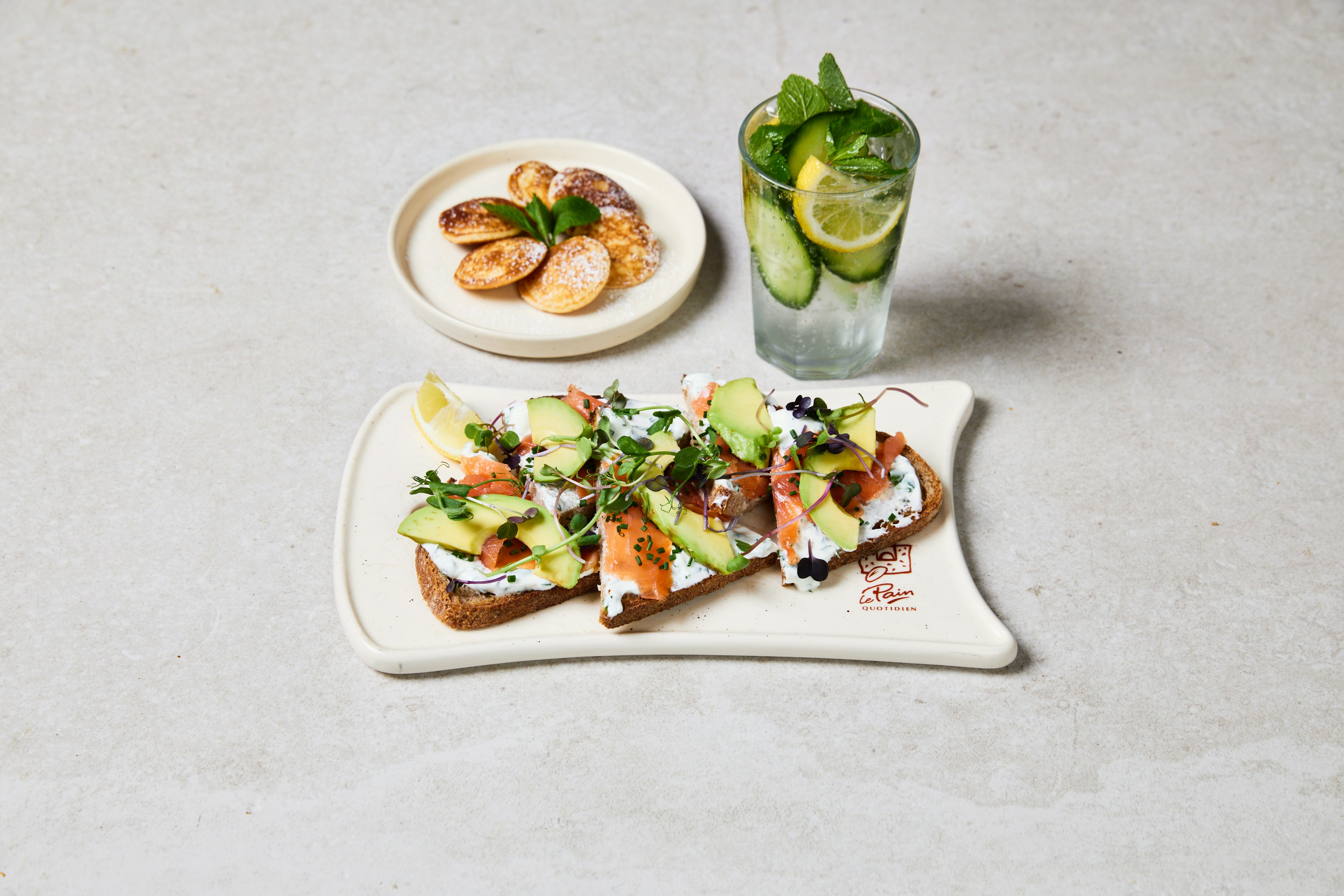 Two slices of toast topped with avocado, smoked salmon, greens, and herbs on a white plate, with a small dish of sliced fruit and a glass of water with cucumber, lemon, and mint.