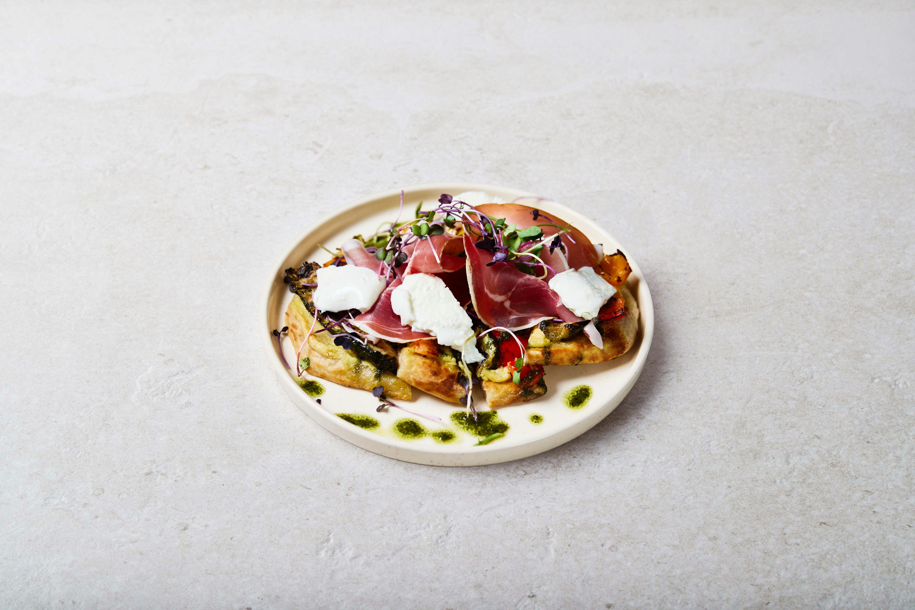 A plated dish featuring slices of prosciutto, dollops of white cheese, microgreens, grilled vegetables, and pesto drizzle, served on a beige plate against a light stone background.