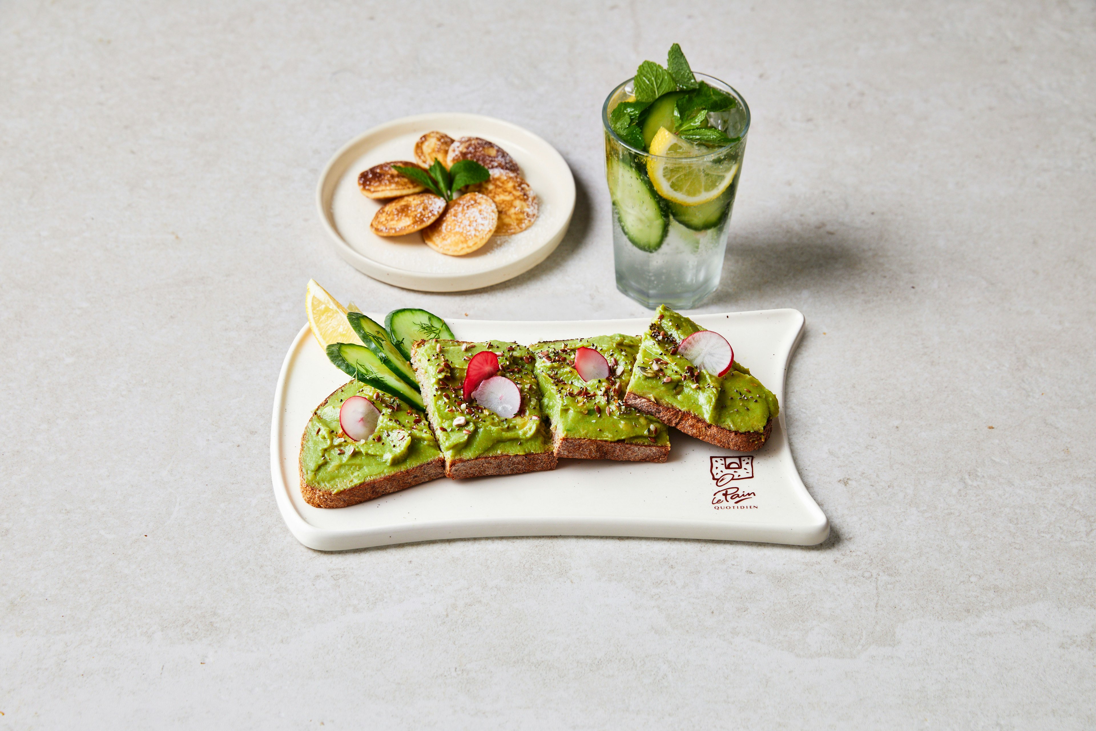 A plate with sliced avocado toast topped with radish and cucumber, a lemon wedge, a side plate of cookies with a green garnish, and a glass of cucumber and mint water on a light surface.