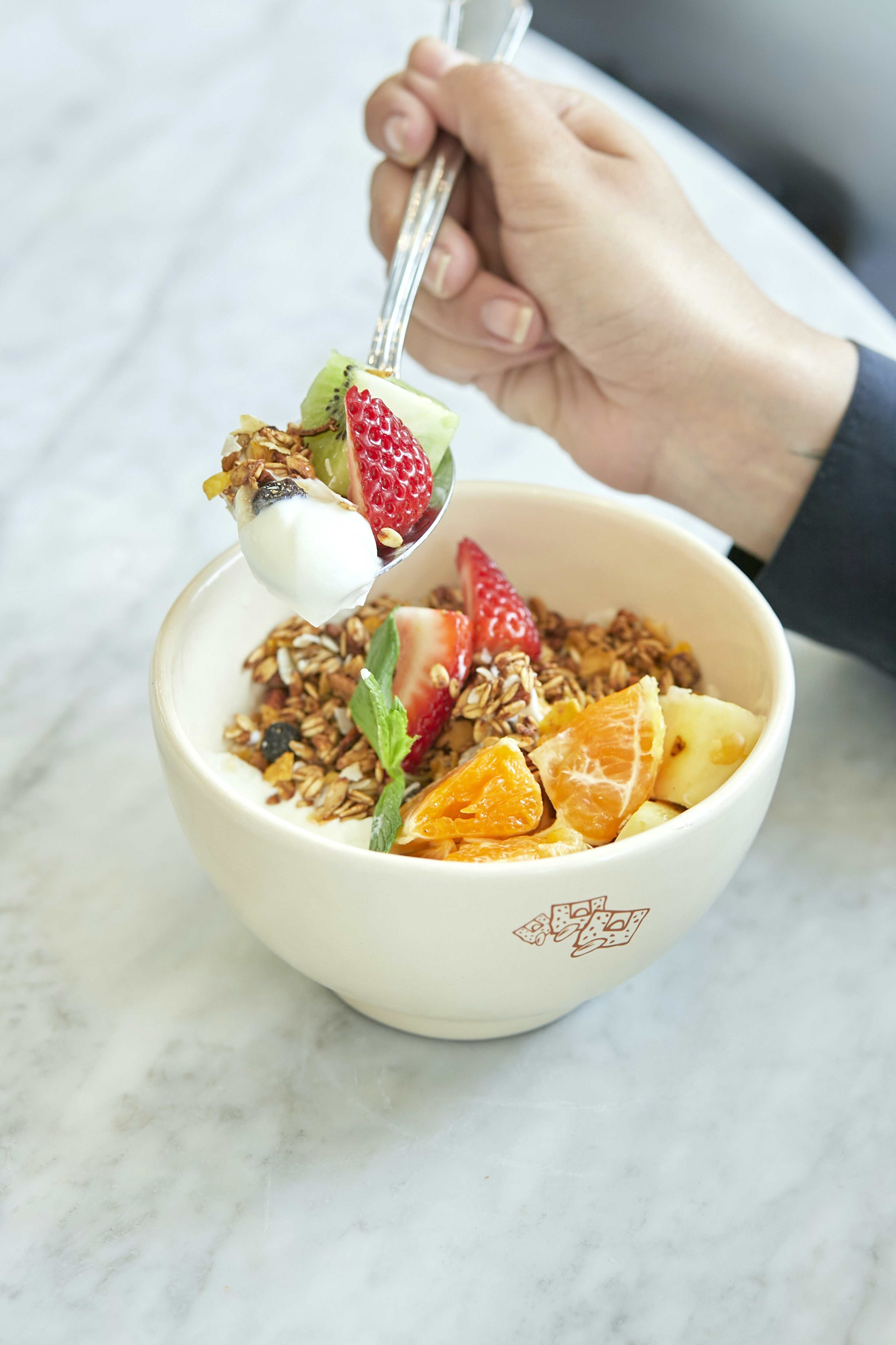 A hand holds a spoonful of yogurt, granola, and fresh fruit over a bowl containing more yogurt, granola, and assorted fruit slices on a marble surface.