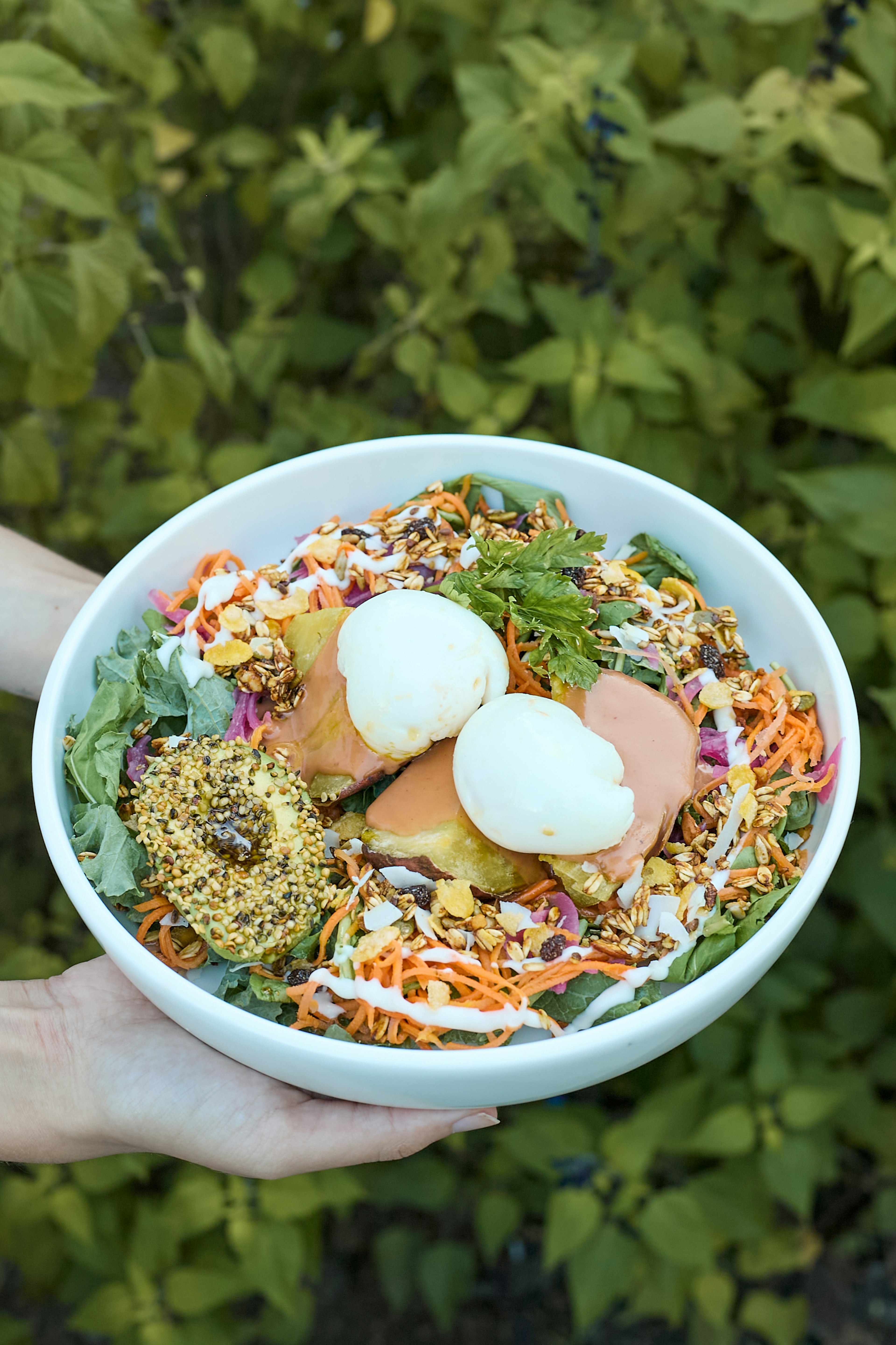 A person holds a white bowl filled with a colorful salad, featuring leafy greens, shredded carrots, seeds, two poached eggs, sauce, and a dollop of grainy mustard, with green foliage in the background.