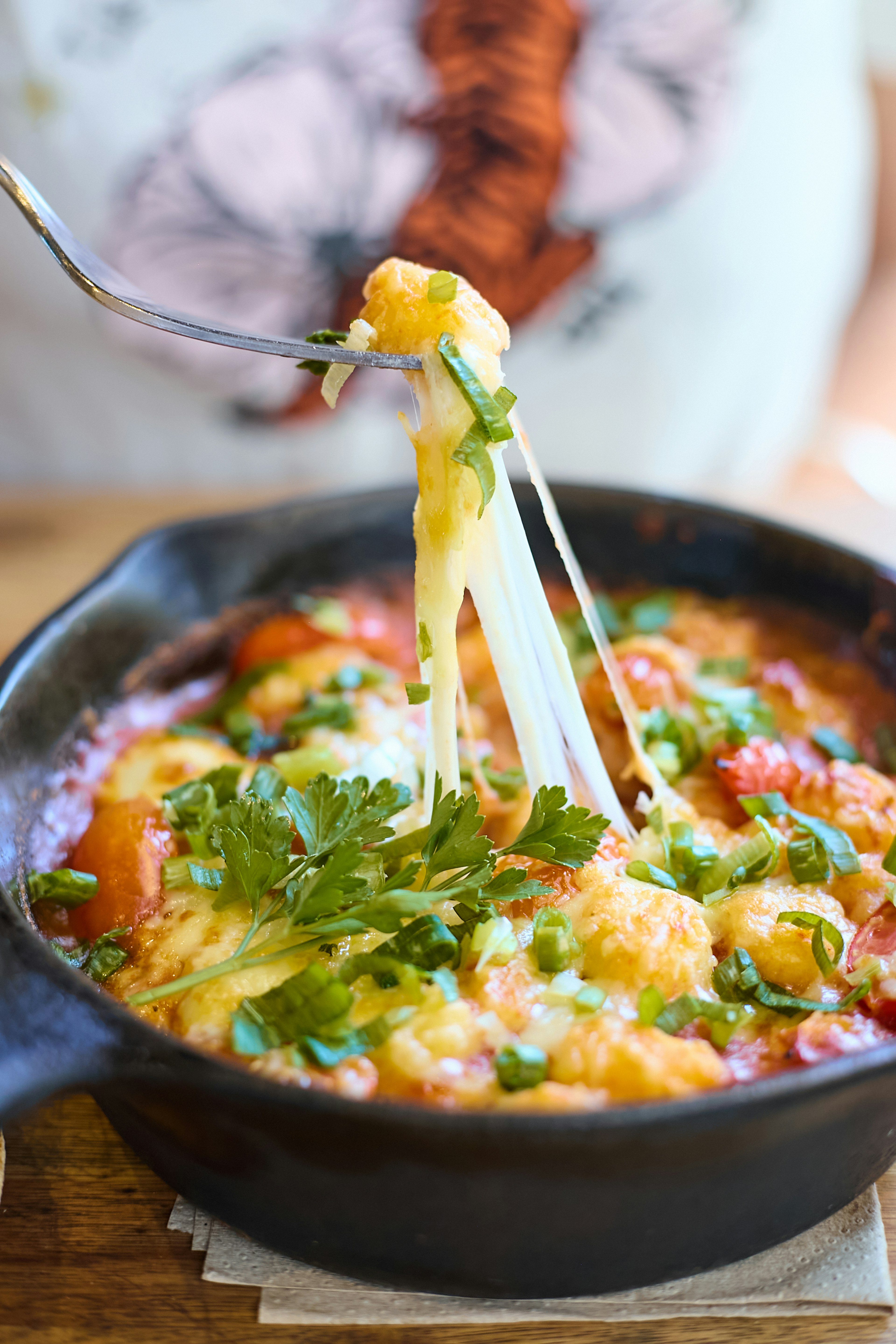 A fork lifts a cheesy, stretchy bite of baked gnocchi out of a cast iron skillet, garnished with fresh herbs. The dish appears hot and freshly cooked, with melted cheese and chopped greens visible.