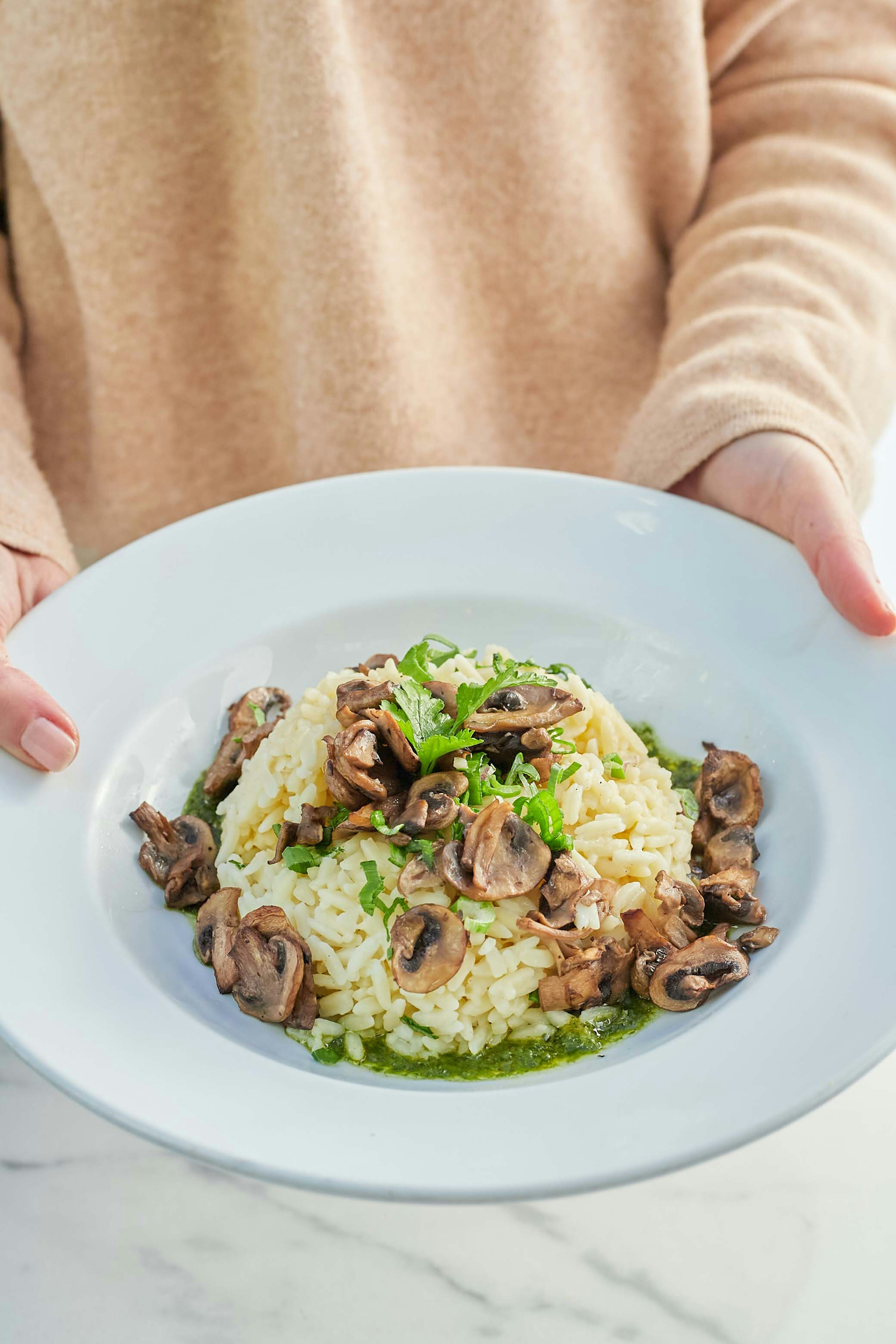 A person in a beige sweater holds a white plate with creamy risotto, topped with sautéed mushrooms and garnished with fresh herbs, over a marble surface.