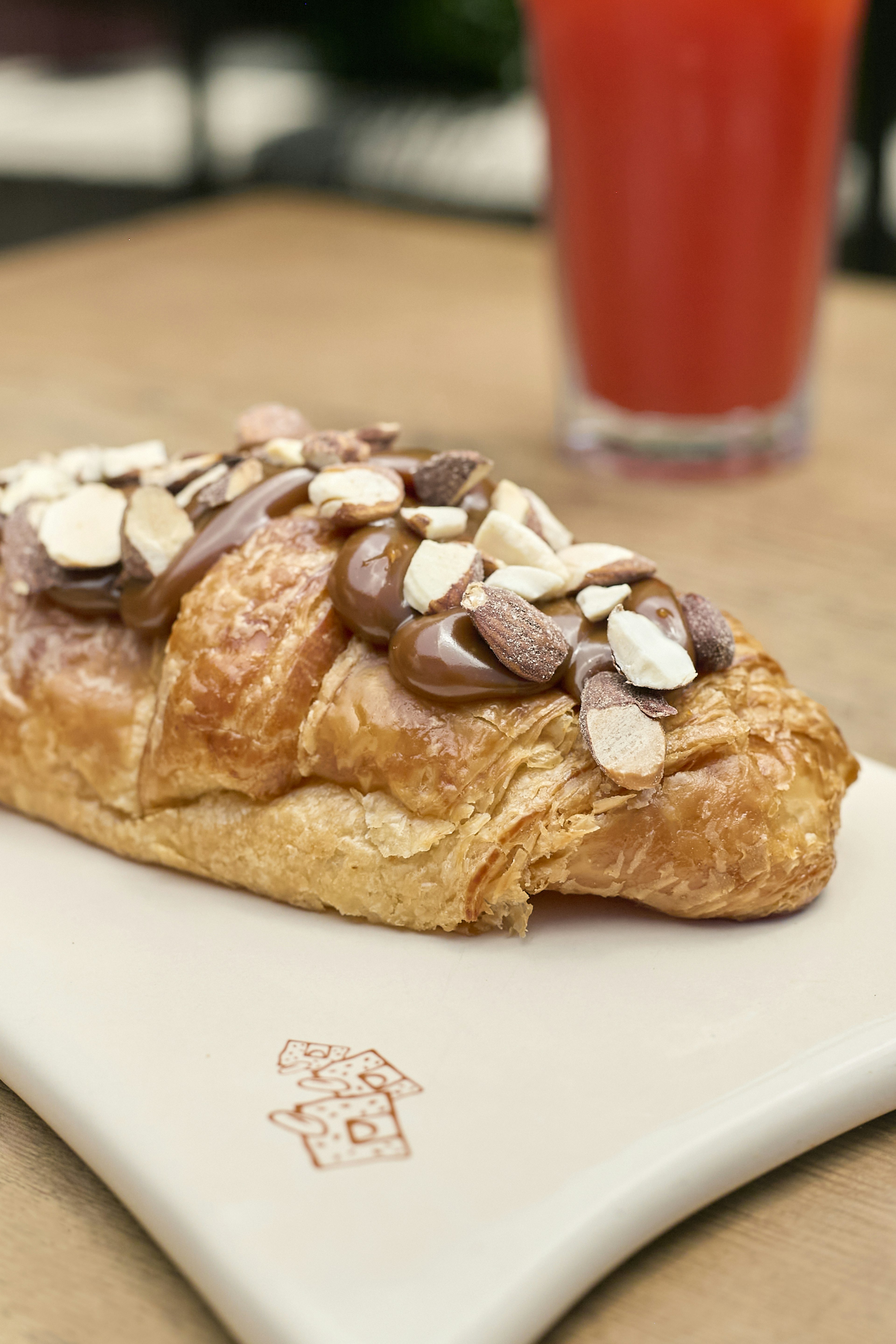 A croissant topped with chocolate spread and sliced almonds sits on a white rectangular plate, with a glass of red juice blurred in the background.