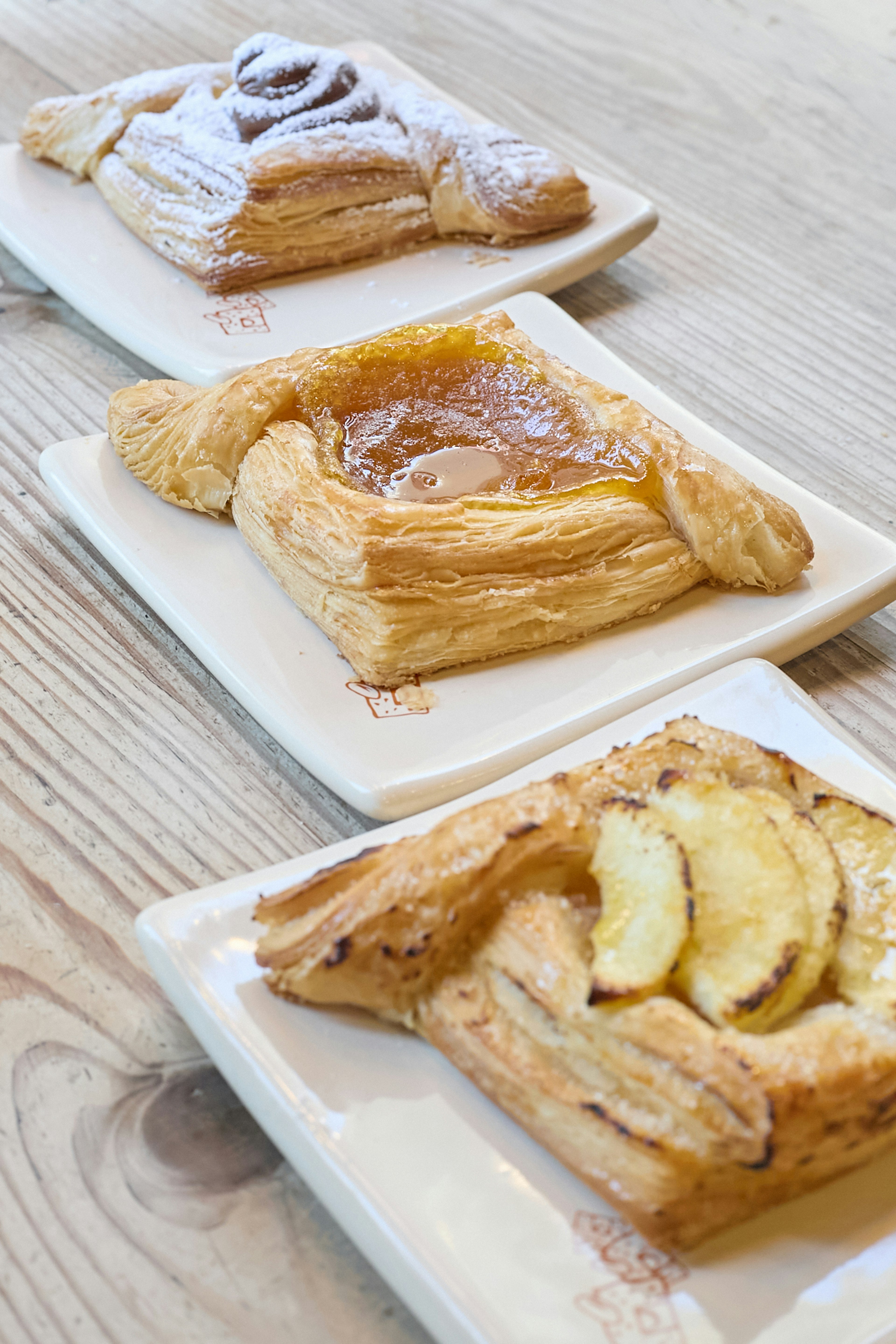 Three square pastries with flaky crusts and various fruit toppings are displayed on white plates, arranged in a row on a light wooden table.