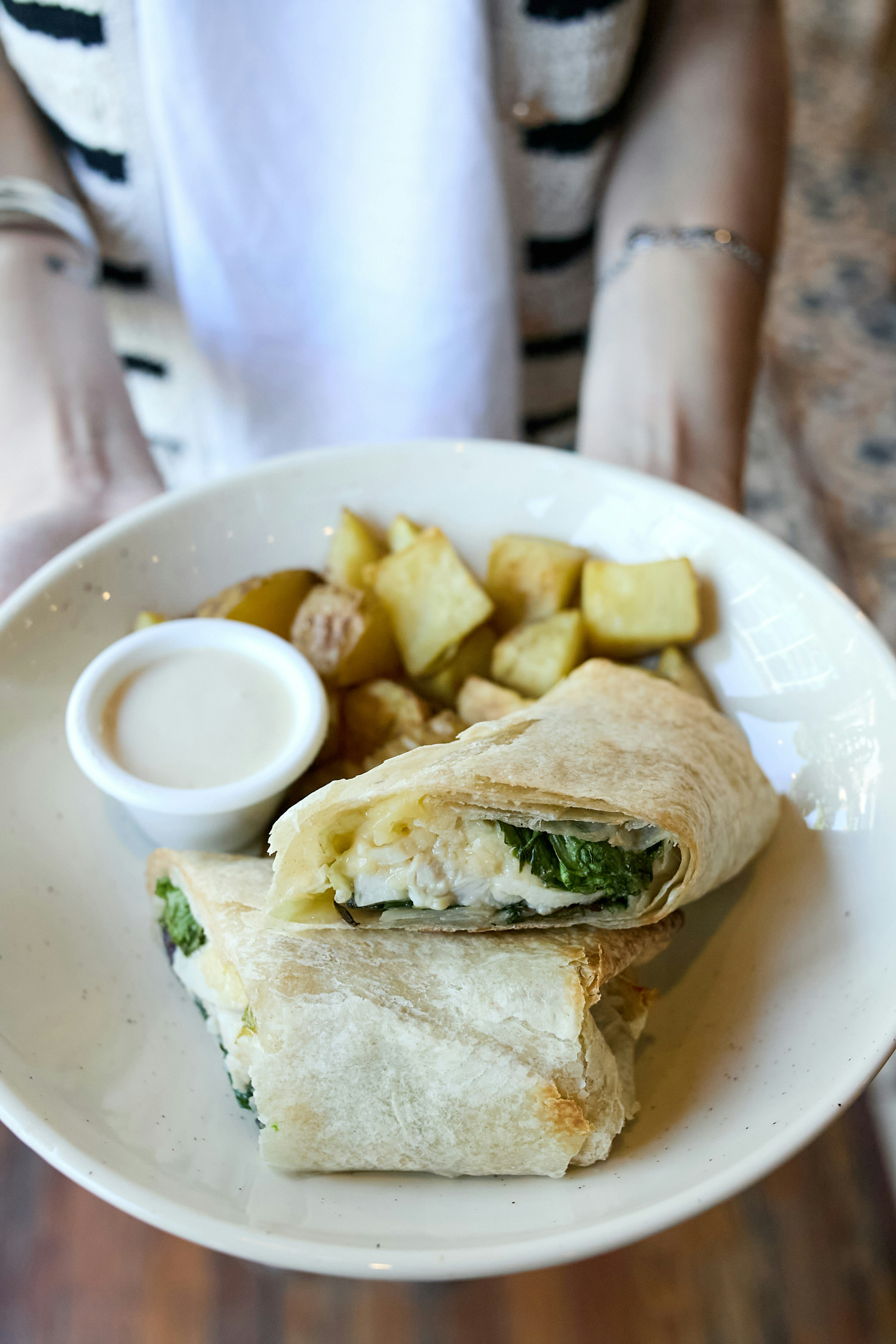 A person holds a plate with a wrap cut in half, filled with greens and cheese, roasted potato cubes, and a small cup of dipping sauce. The person wears a striped cardigan.