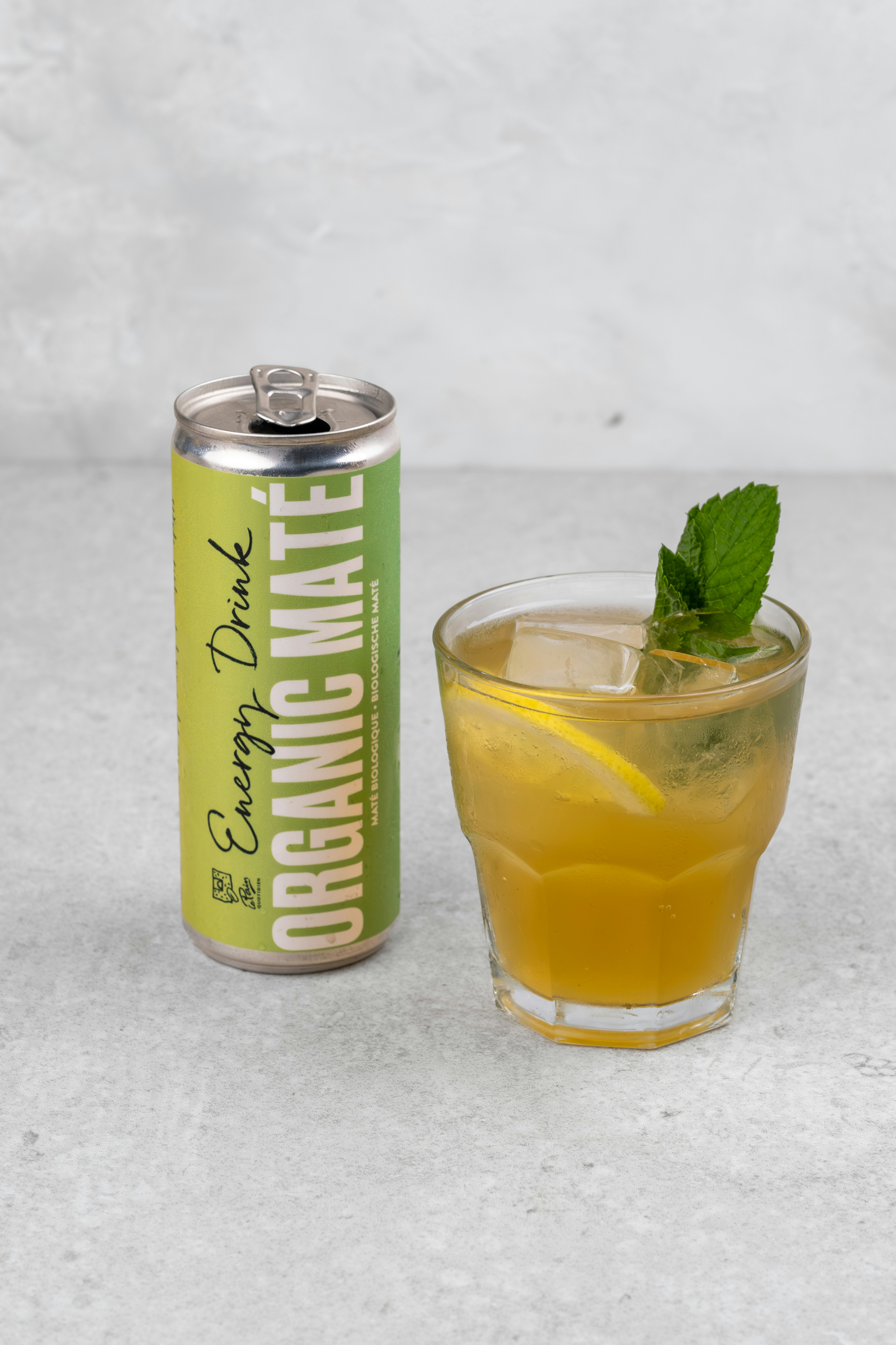 A can labeled Energy Drink Organic Mate stands next to a glass filled with iced yellow-green drink, garnished with a lemon slice and mint, on a light gray surface.
