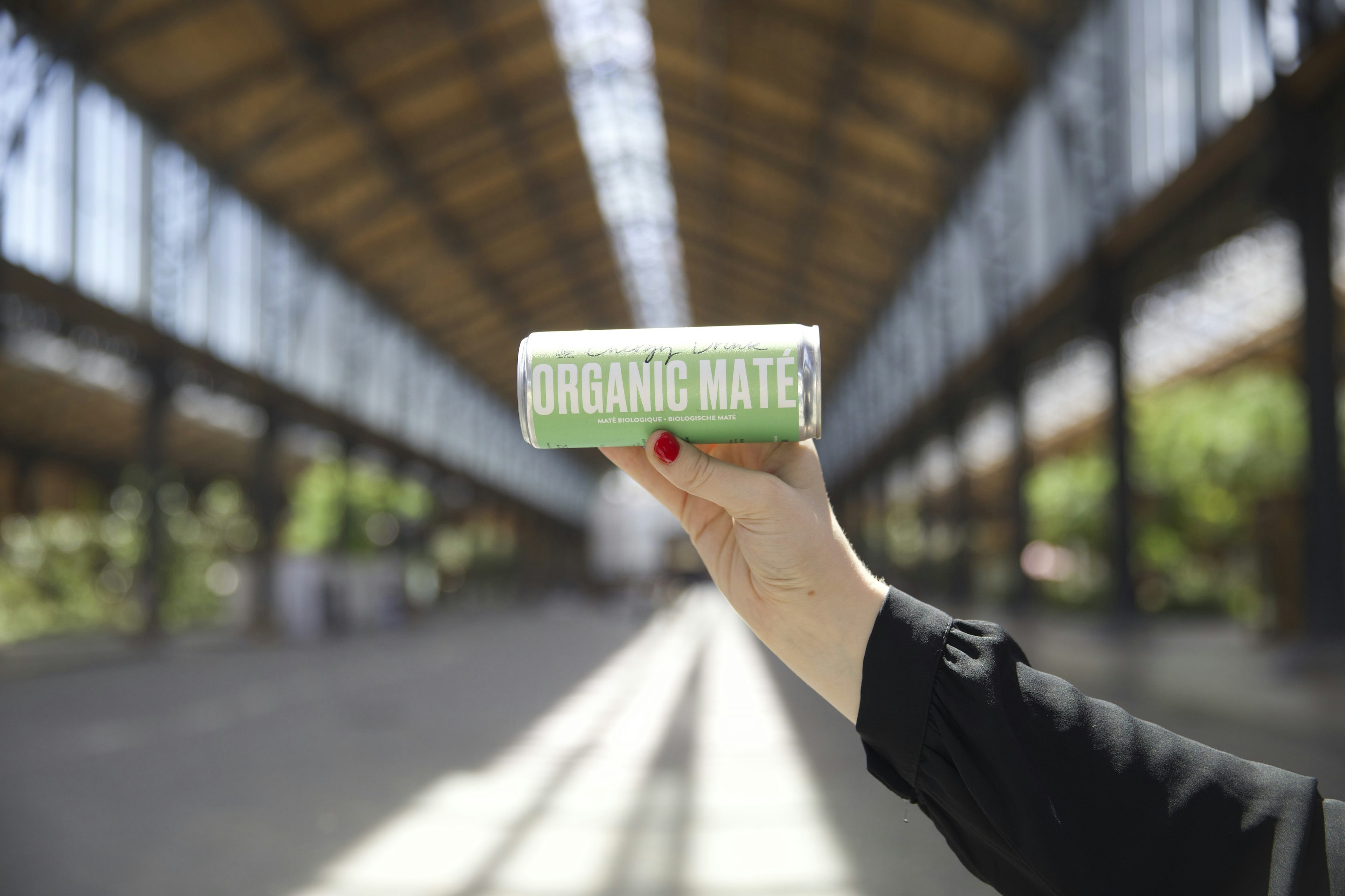 A hand with red nail polish holds a can labeled ORGANIC MATE inside a spacious, sunlit building with a high glass ceiling and metal framework.