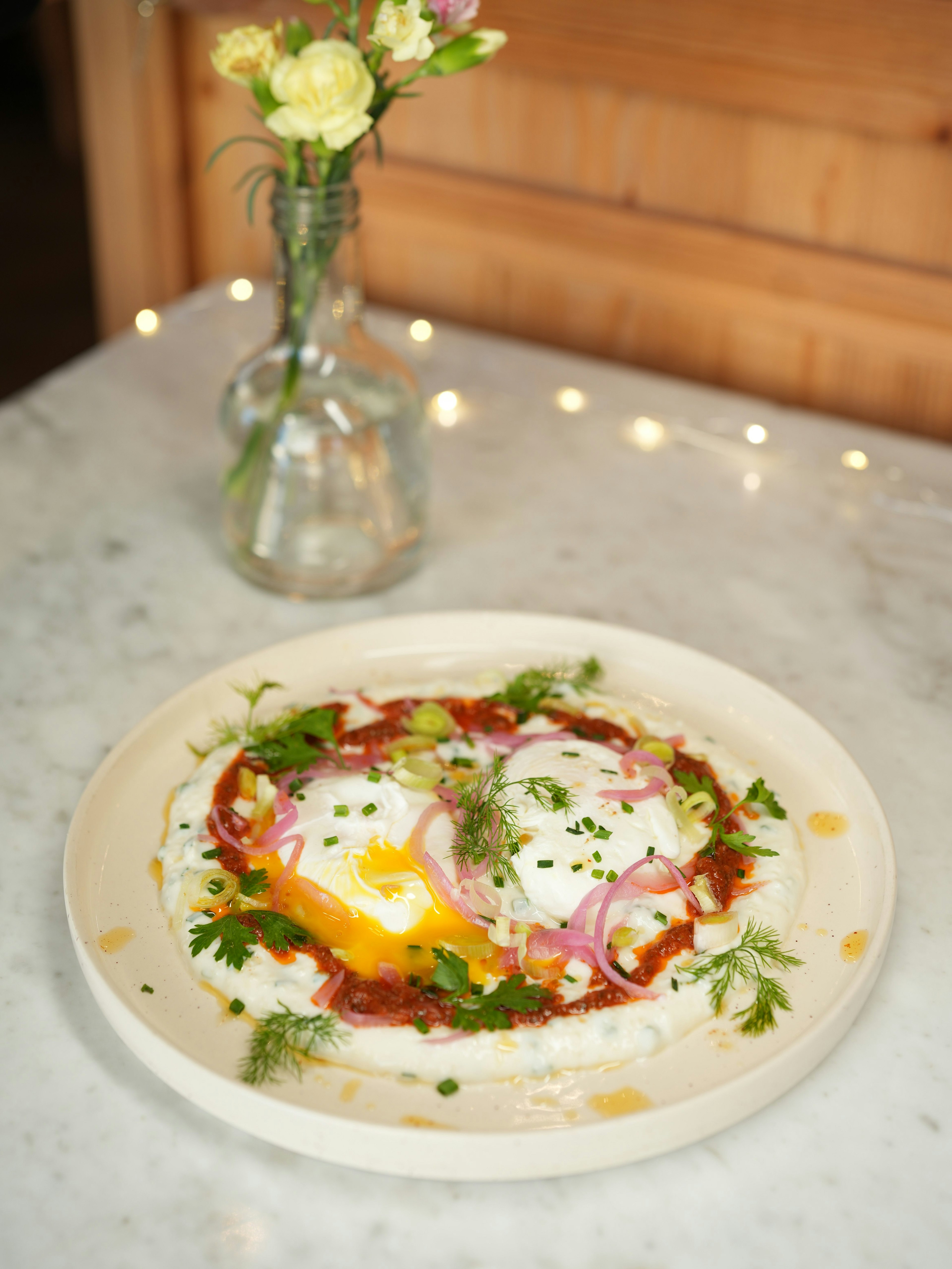 A plate with two sunny-side-up eggs on yogurt, garnished with herbs, red onions, and spices sits on a marble table. A glass vase with small yellow flowers is in the background.