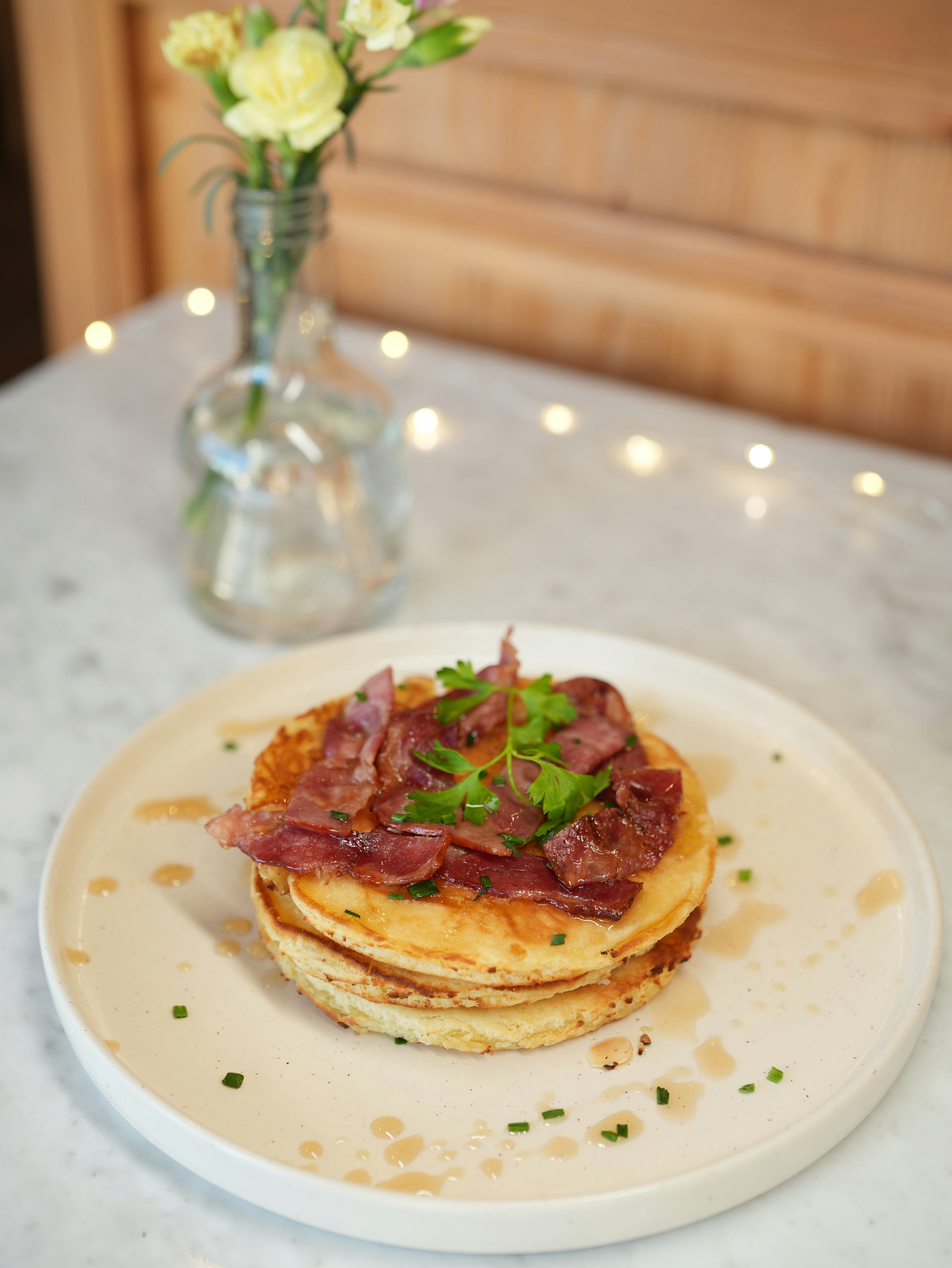 A stack of pancakes topped with crispy bacon and fresh herbs, drizzled with syrup, sits on a white plate. A glass vase with yellow flowers is in the background on a marble table.