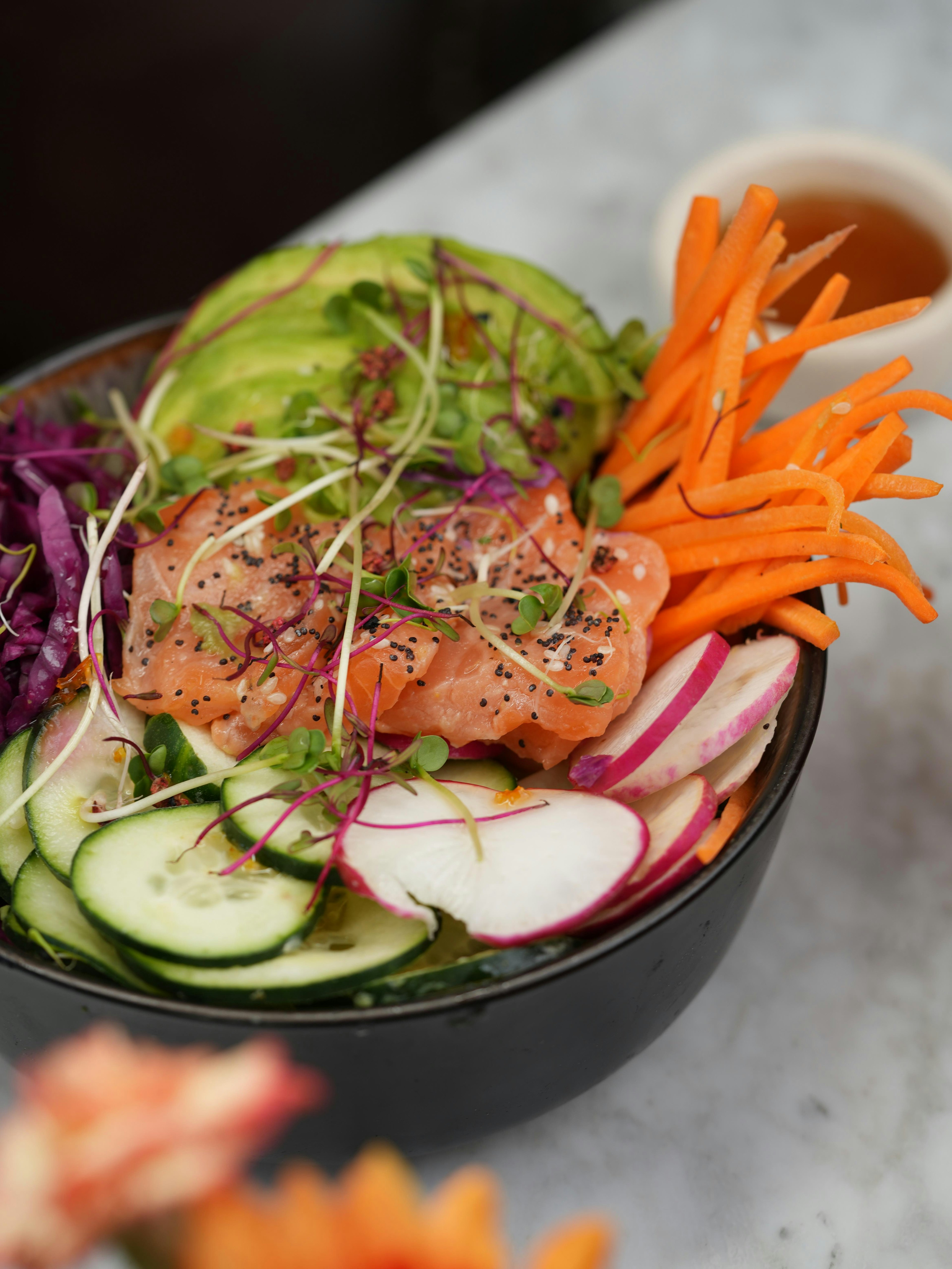 A poke bowl with sliced salmon, avocado, shredded carrots, cucumber, radish, red cabbage, and microgreens, served in a black bowl on a marble surface with a sauce cup nearby.