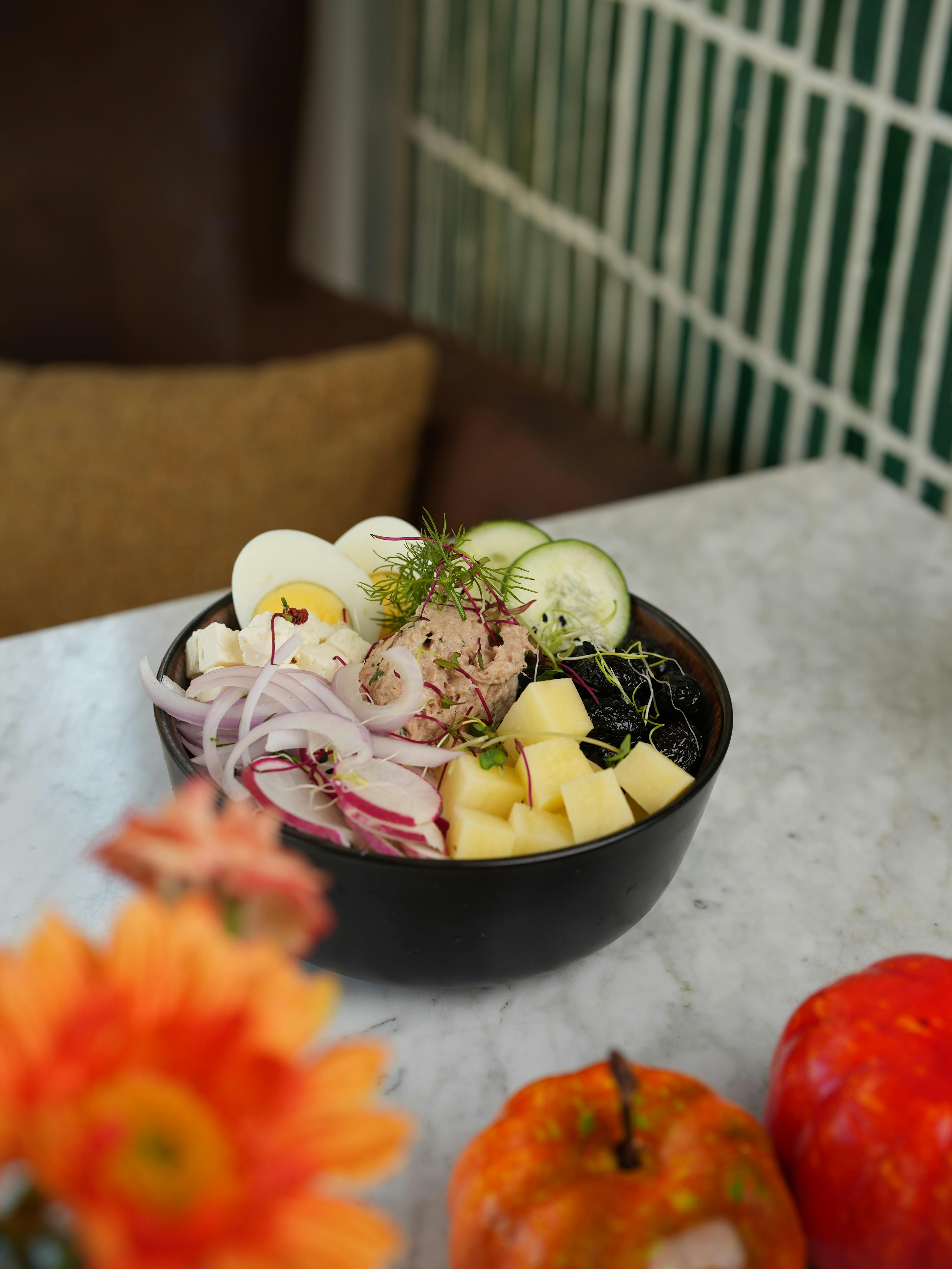 A black bowl with sliced egg, cucumber, cheese cubes, red onion, olives, tuna salad, and herbs sits on a marble table, with flowers and fruit in the foreground and a green-tiled wall in the background.