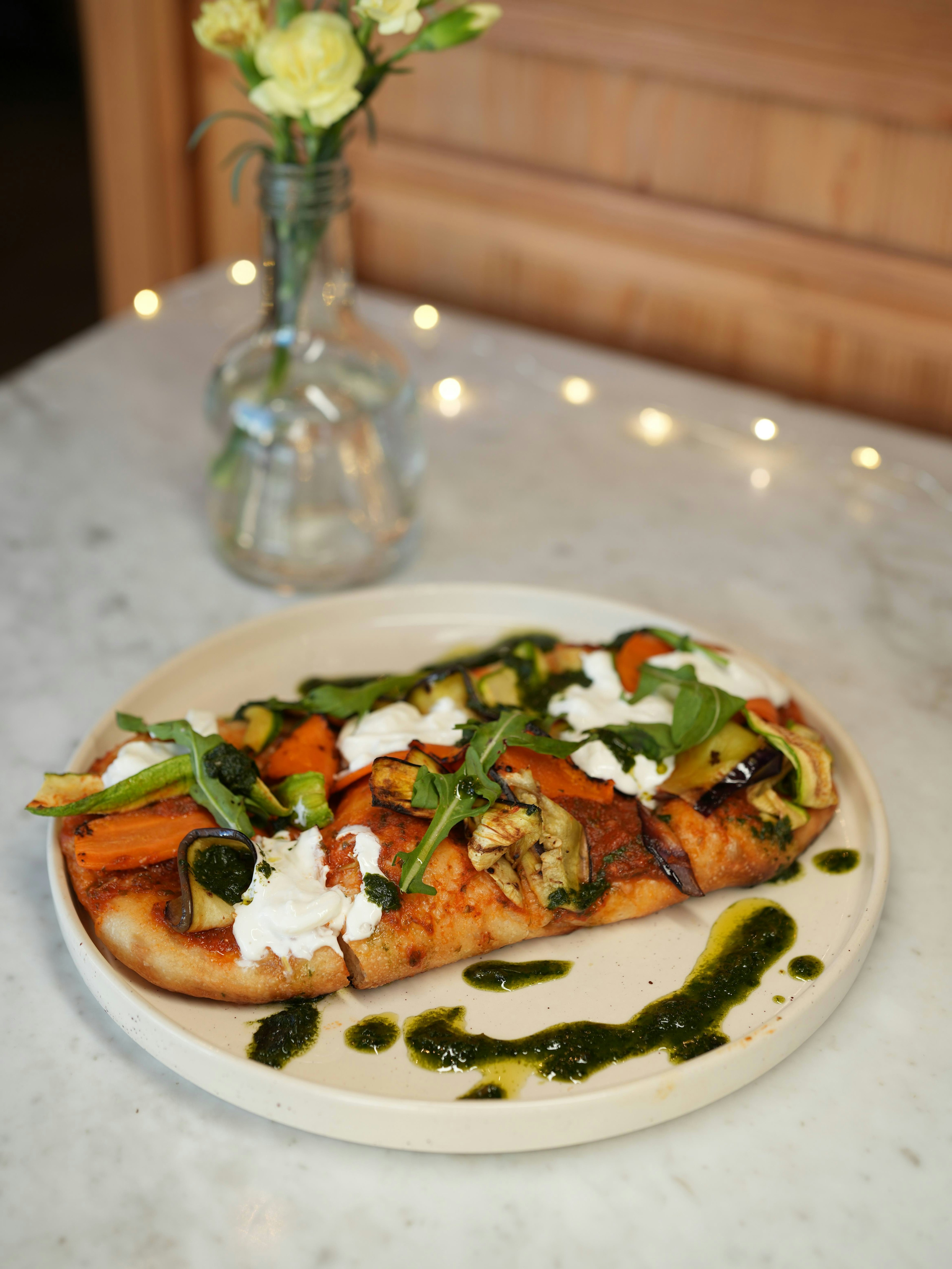 A flatbread topped with roasted vegetables, dollops of cheese, and green sauce is served on a white plate. A small vase with yellow flowers sits in the background on a marble table.