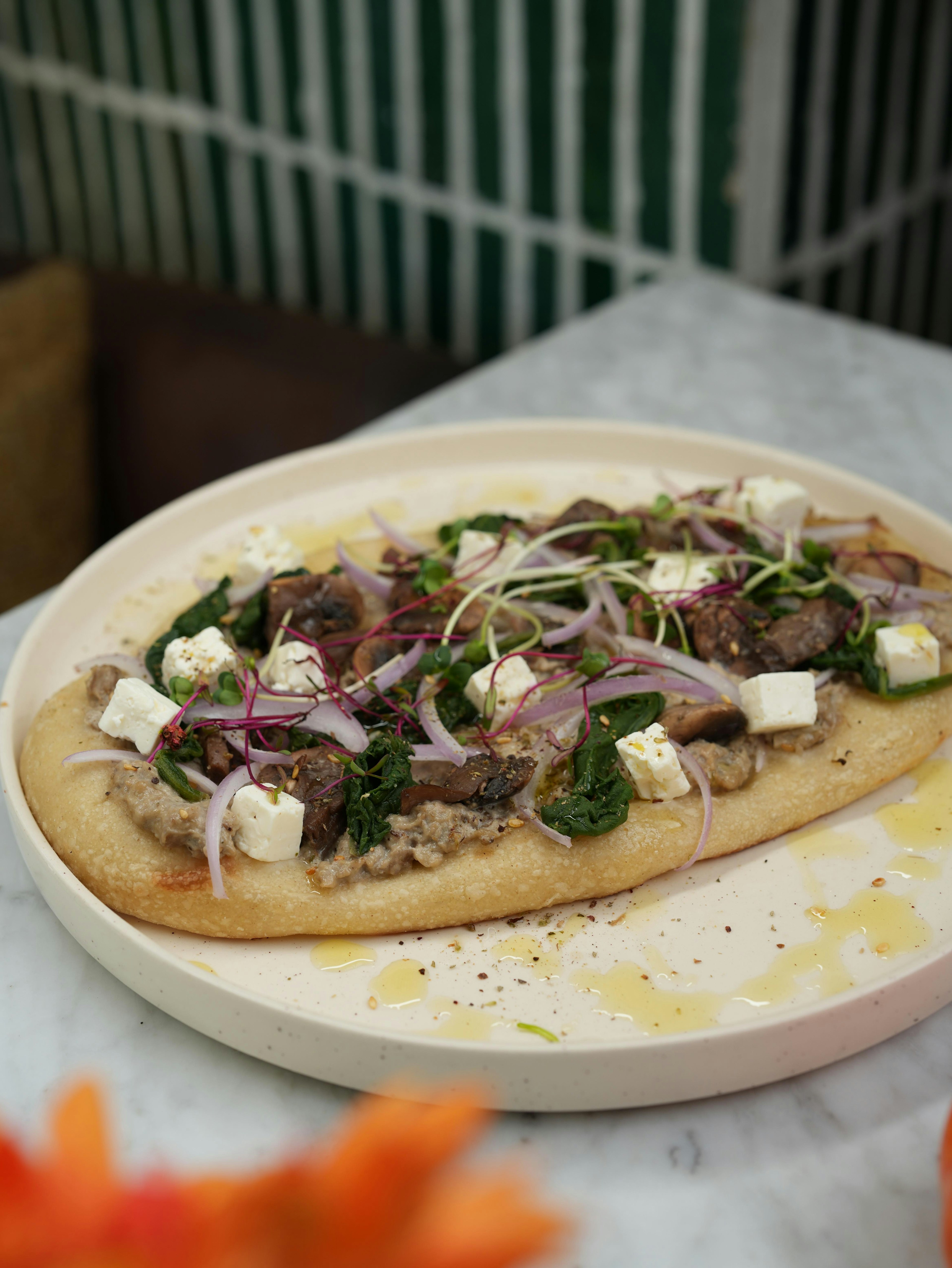 Flatbread topped with chunks of feta cheese, red onions, greens, microgreens, and pieces of meat, served on a round cream-colored plate with a drizzle of oil, on a marble table.