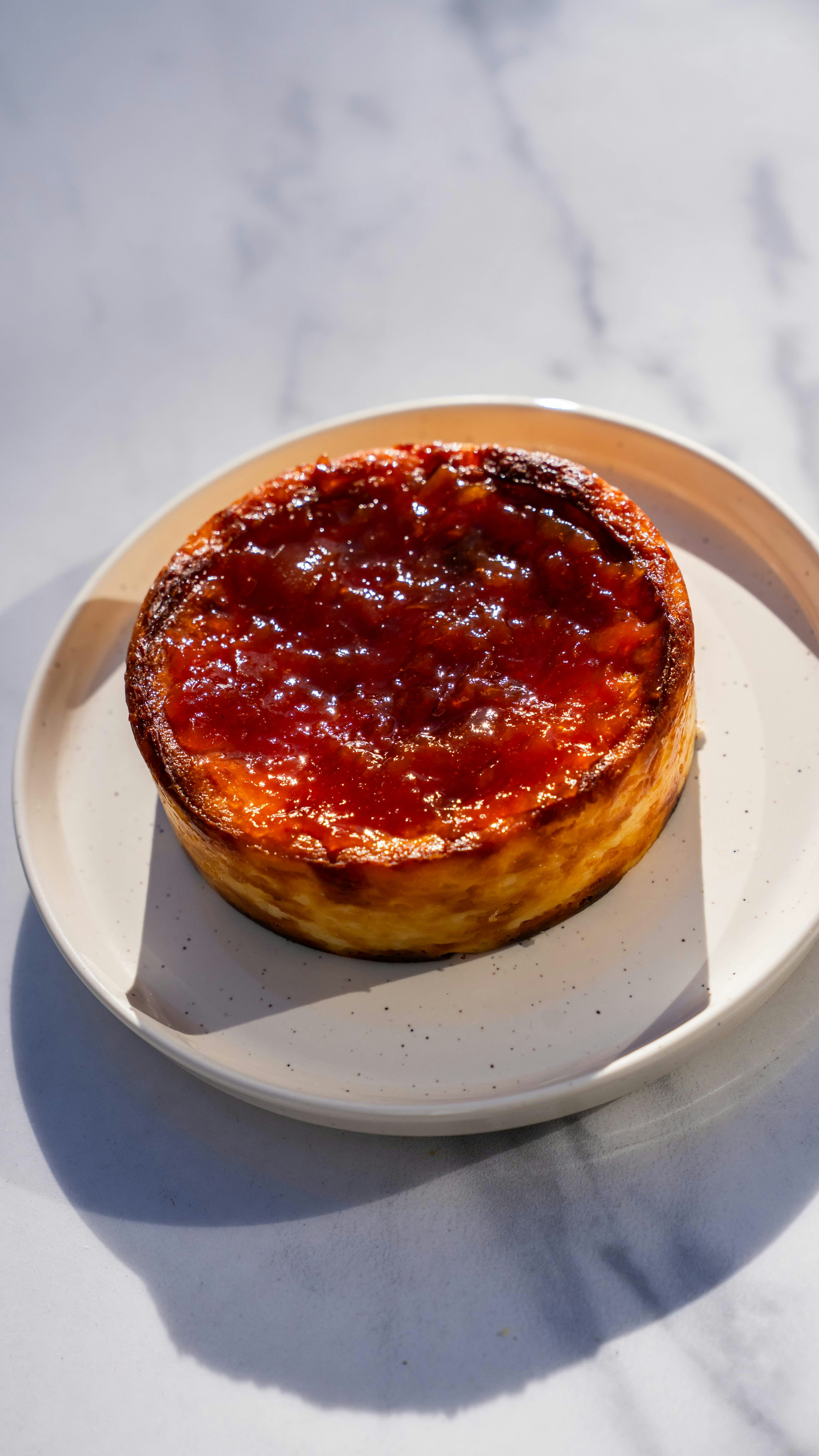 Una tarta de queso vasca redonda, dorada al horno y con la superficie caramelizada, descansa sobre un plato blanco, que a su vez se encuentra sobre una superficie de mármol, iluminada por la luz natural del sol.
