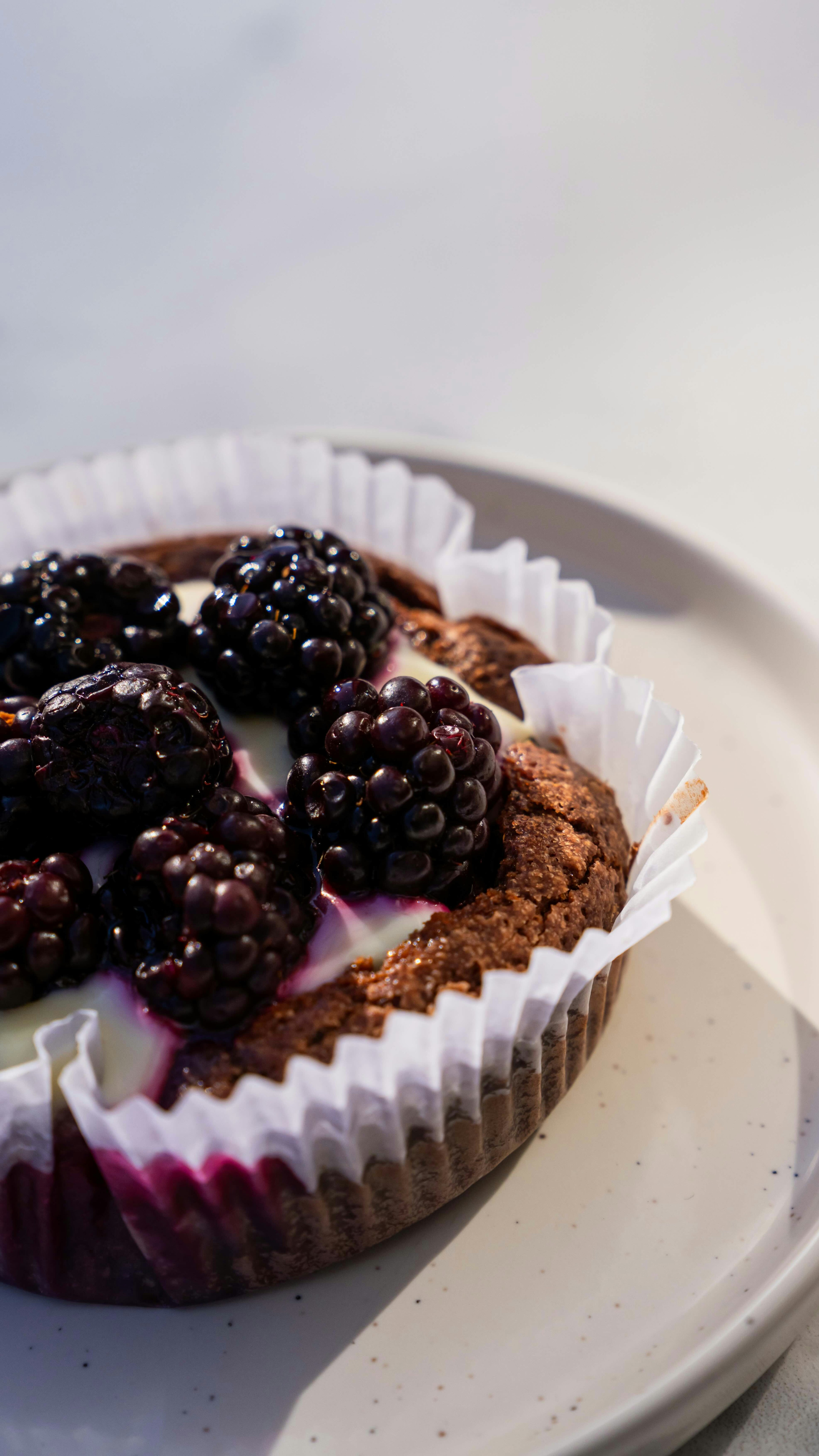 Un muffin de chocolate con moras y glaseado blanco, envuelto en papel, reposa sobre un plato blanco con un fondo claro.