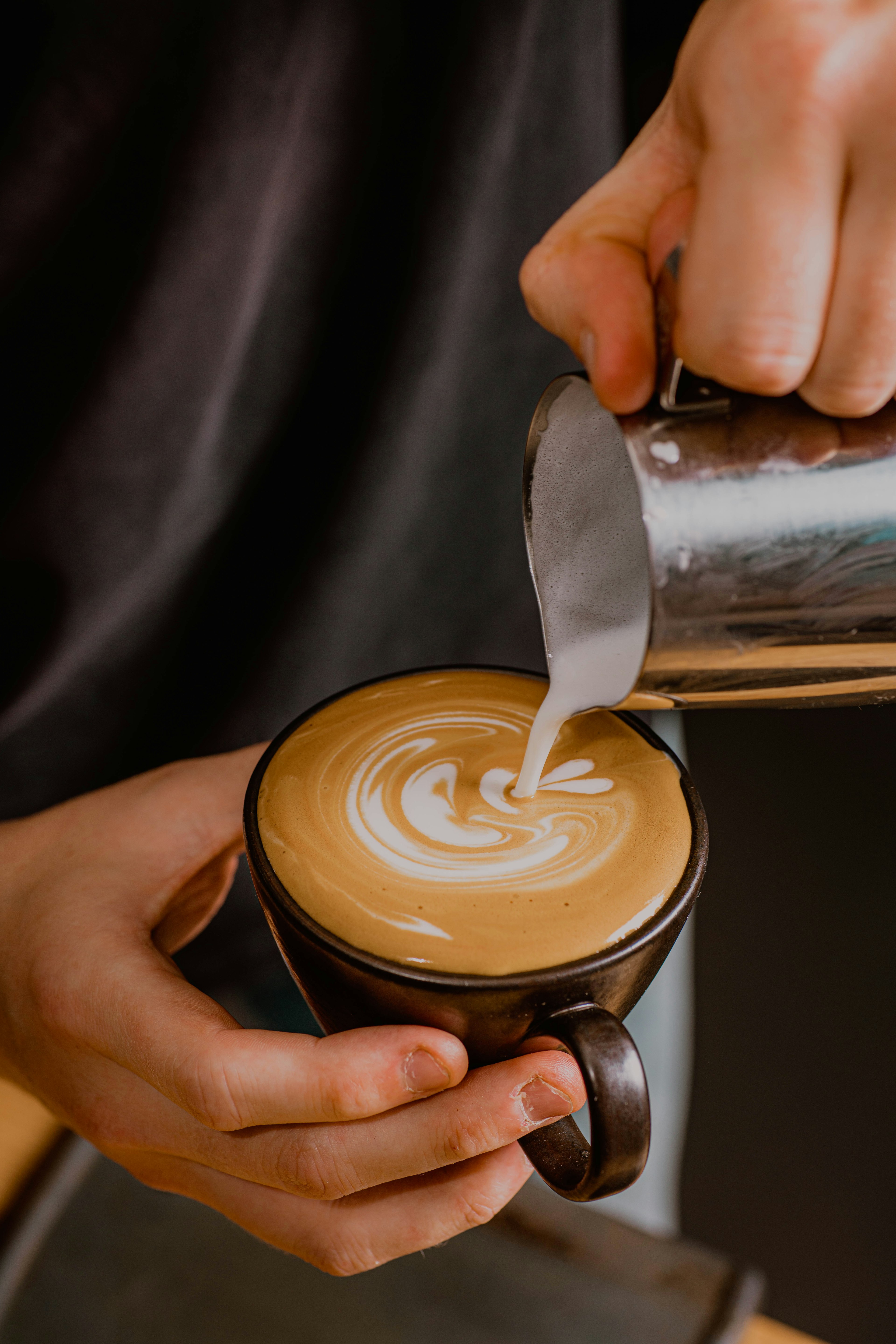 A barista pours steamed milk from a steel jug into a dark ceramic cup, creating a swirling latte art pattern.