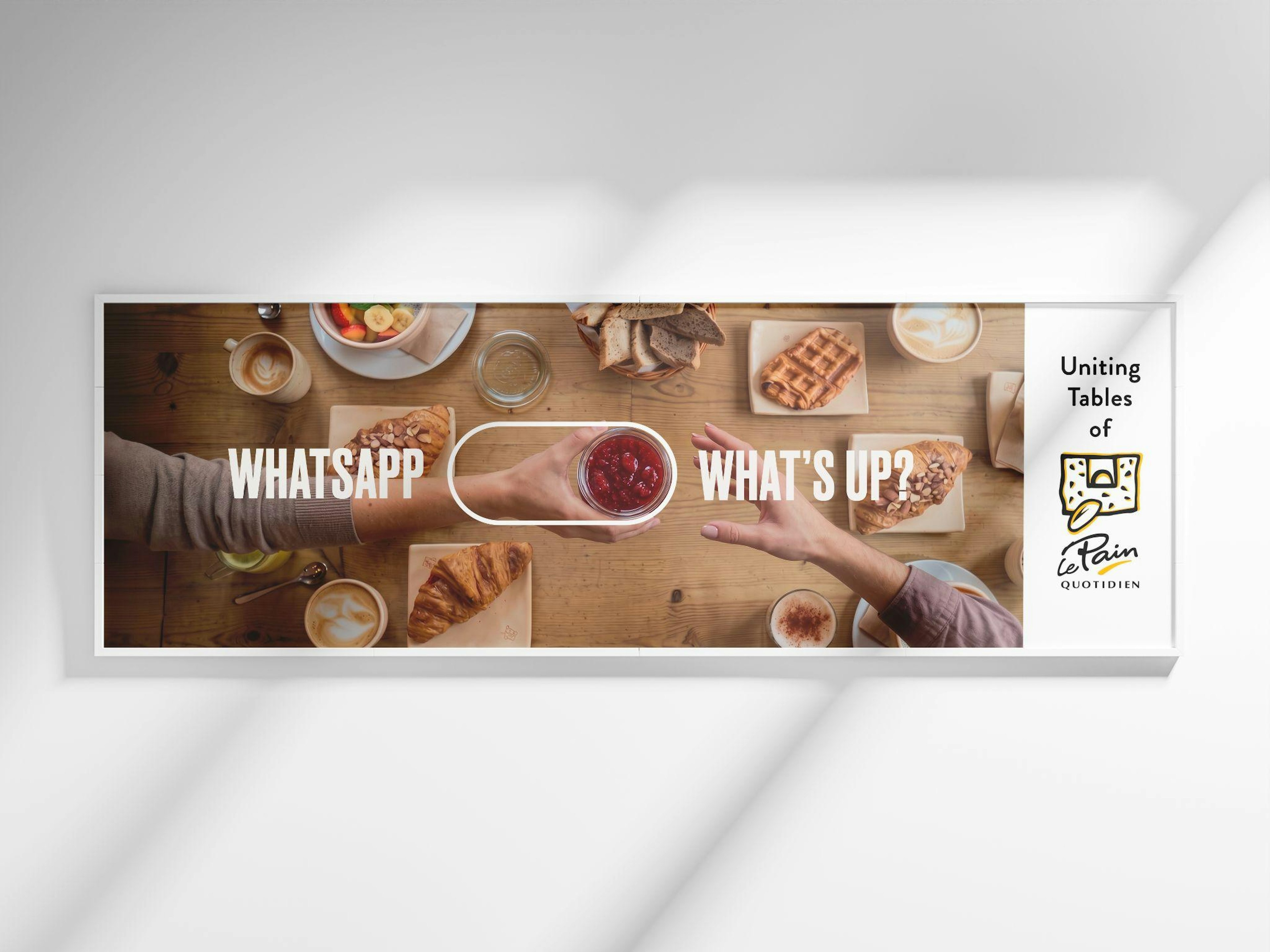 Le Pain Quotidien “Uniting Tables” banner: a hand passes a jar of jam across a communal table of pastries and coffee, with the text “WhatsApp → What’s up?”