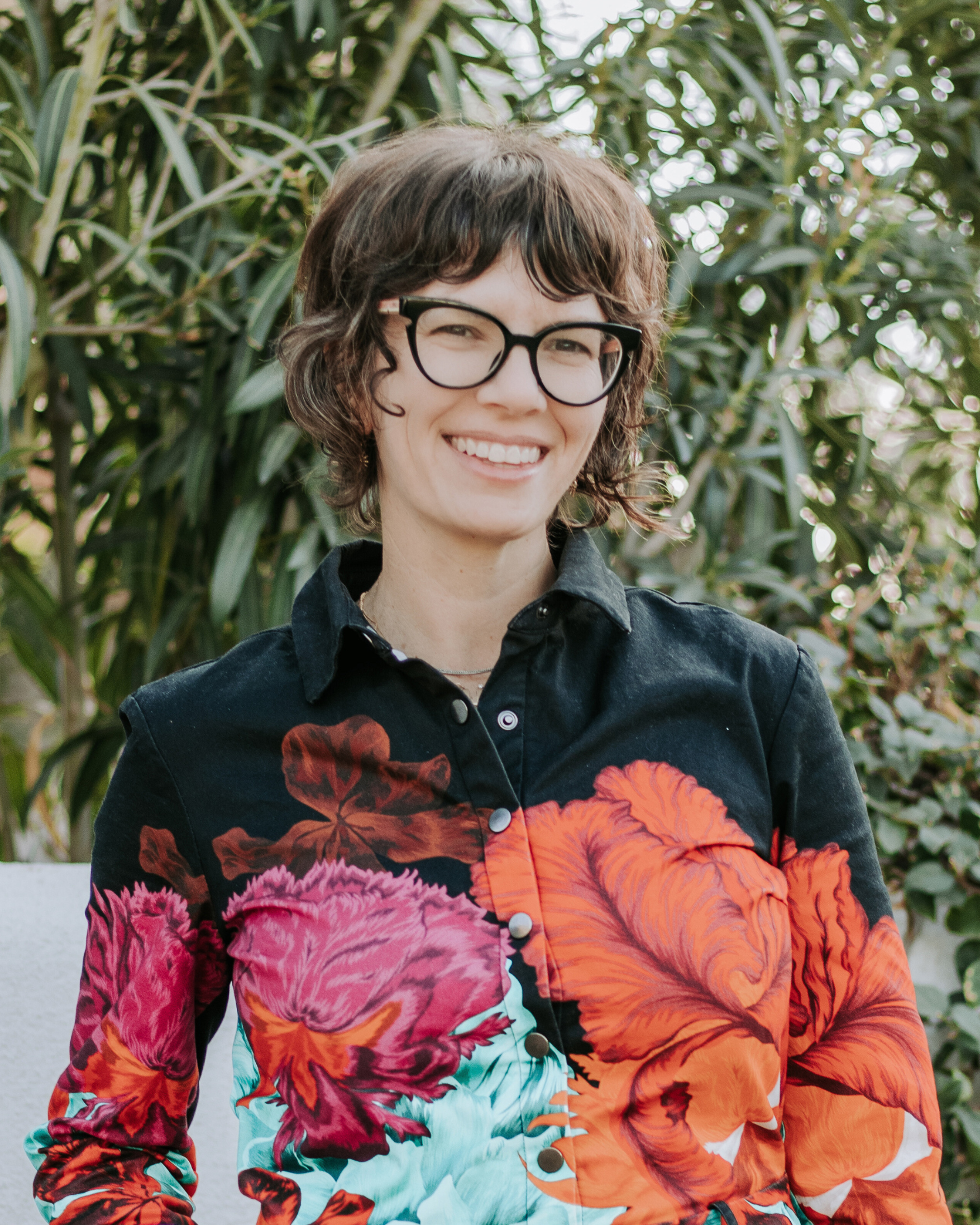 Liz Vogel Executive Director - A woman with short brown hair wearing black-rimmed glasses and a black, long-sleeved collared shirt with flowers on it, standing outside with plants in the background