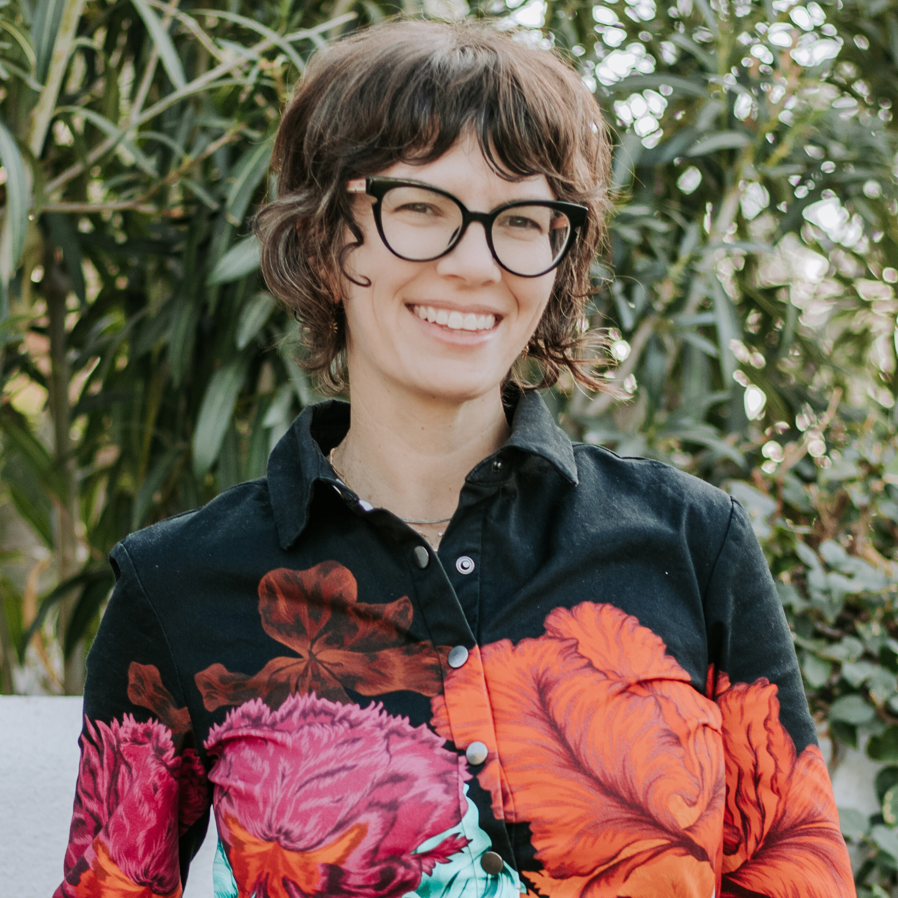 Liz Vogel Executive Director - A woman with short brown hair wearing black-rimmed glasses and a black, long-sleeved collared shirt with flowers on it, standing outside with plants in the background